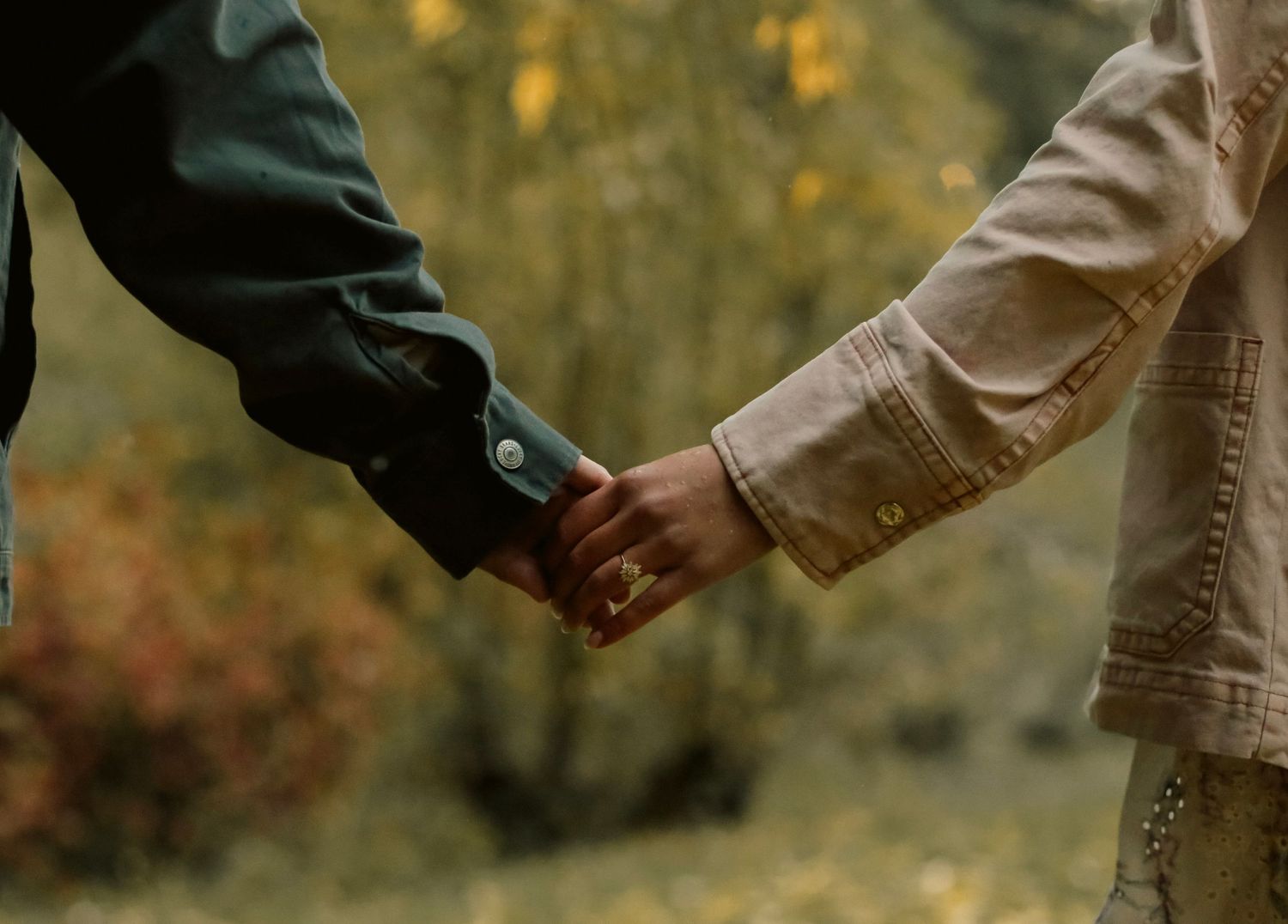 Couple holding hands in forest setting with engagement rings.