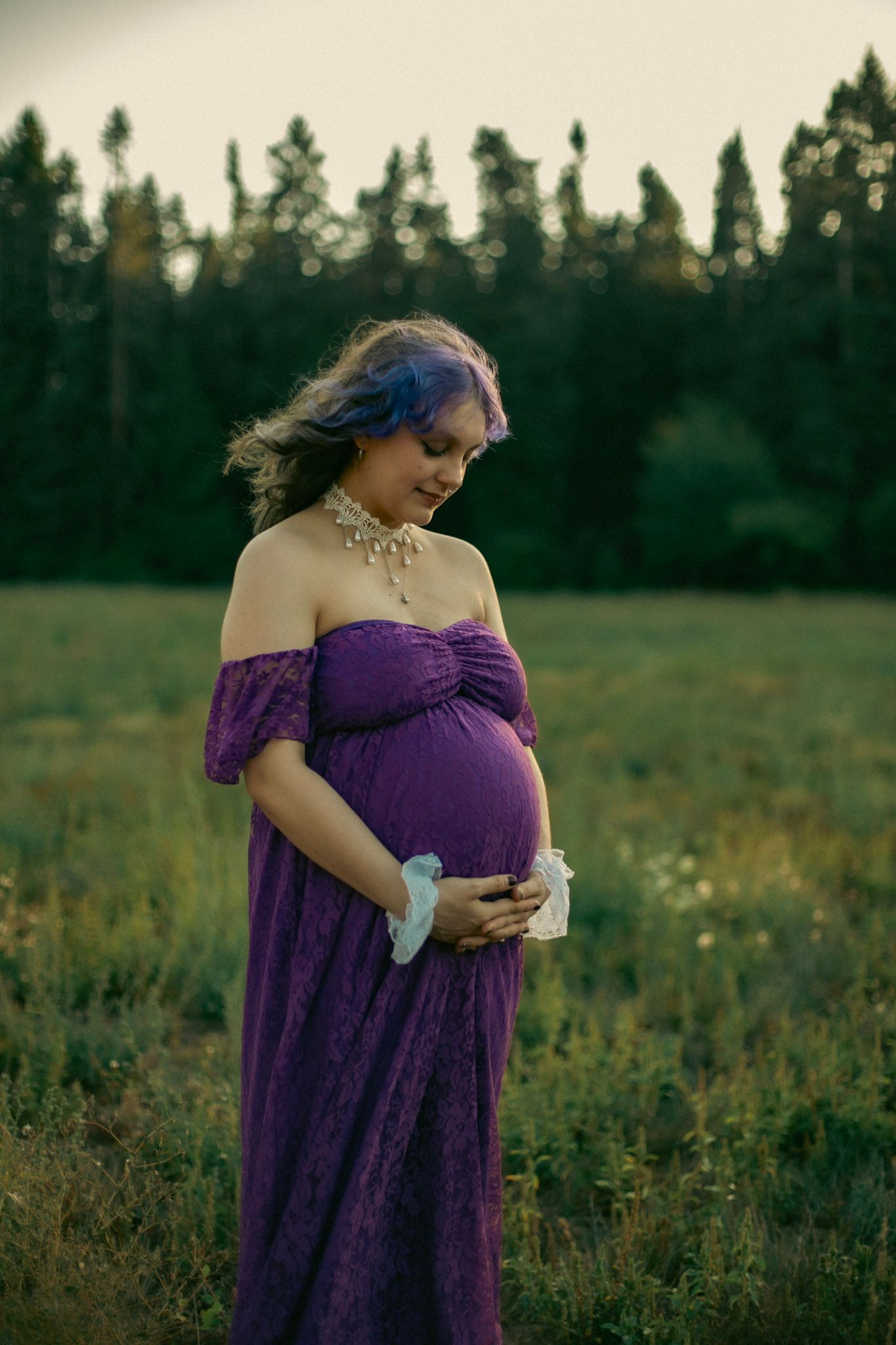Pregnant woman poses in a long purple dress for a maternity photoshoot with a mood treeline background.