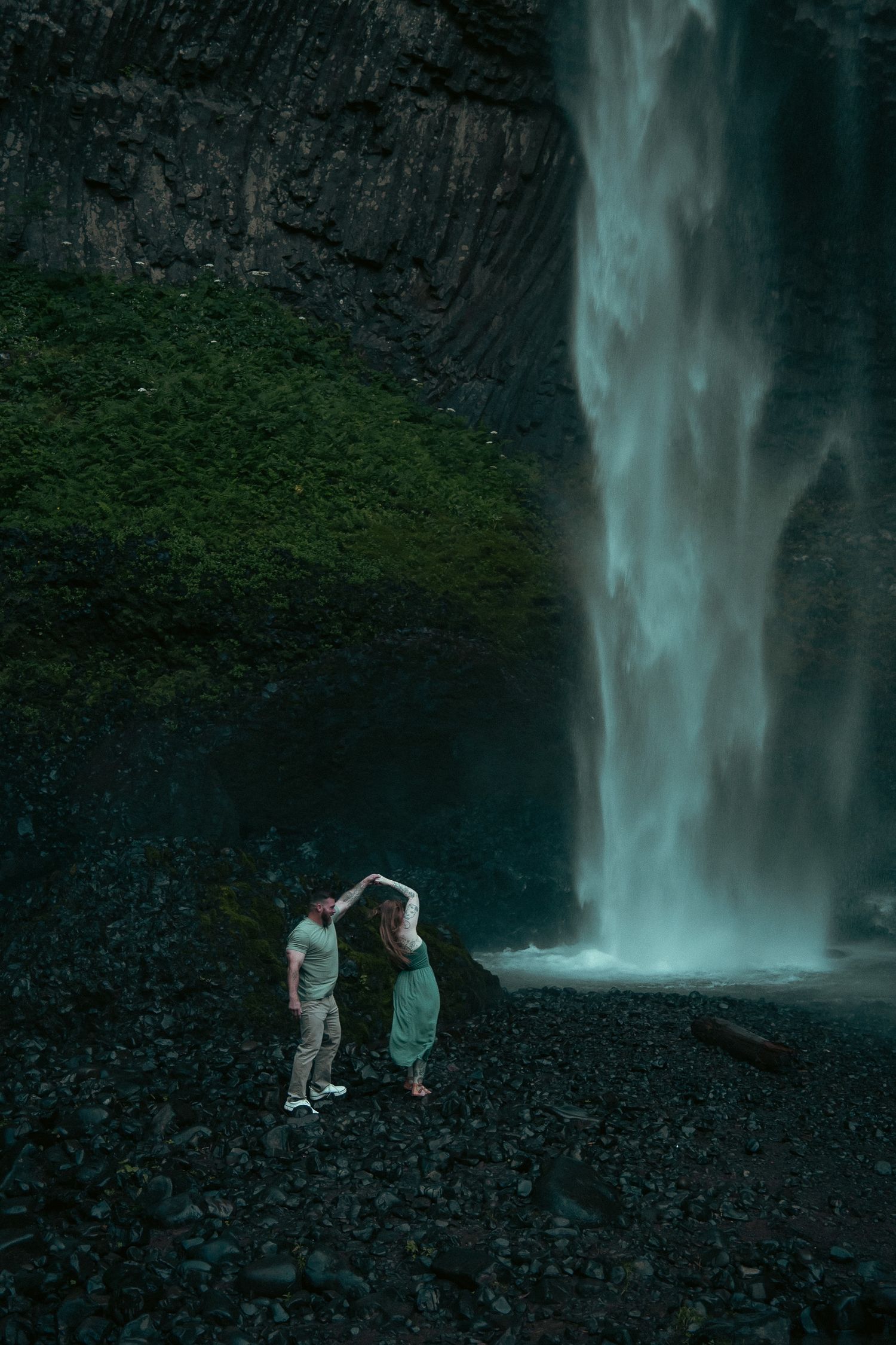 A couple holding hands and twirling in front of a blue waterfall amongst dark, moody rocks for an engagement photoshoot