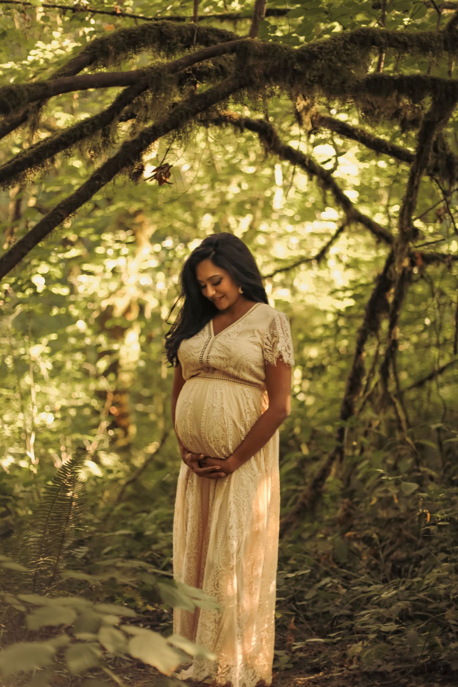 A woman in a long white dress holding her pregnant belly and looking down while standing in a lush, mossy, green forest.