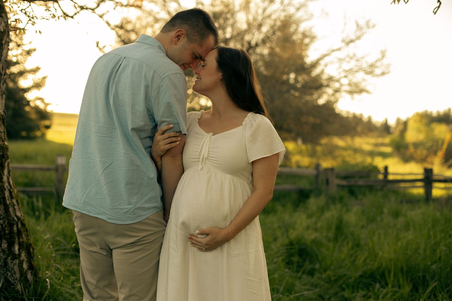 A couple stand facing opposite directions in a green, sunny, country field, looking into each other's eyes and posing for maternity photos