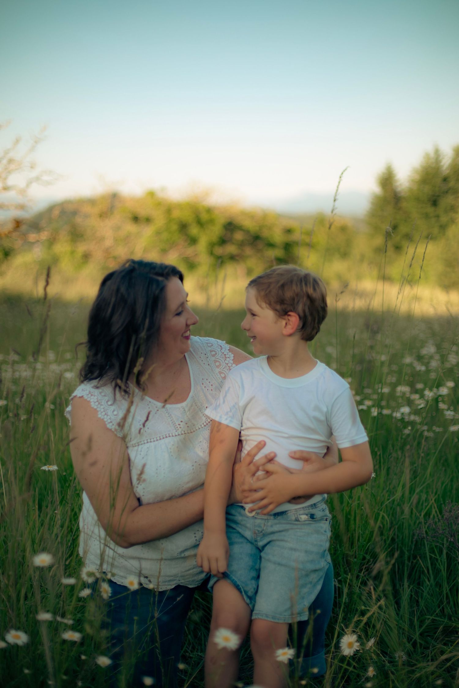A mother and son sitting in a green field with white flowers looking at each other lovingly.