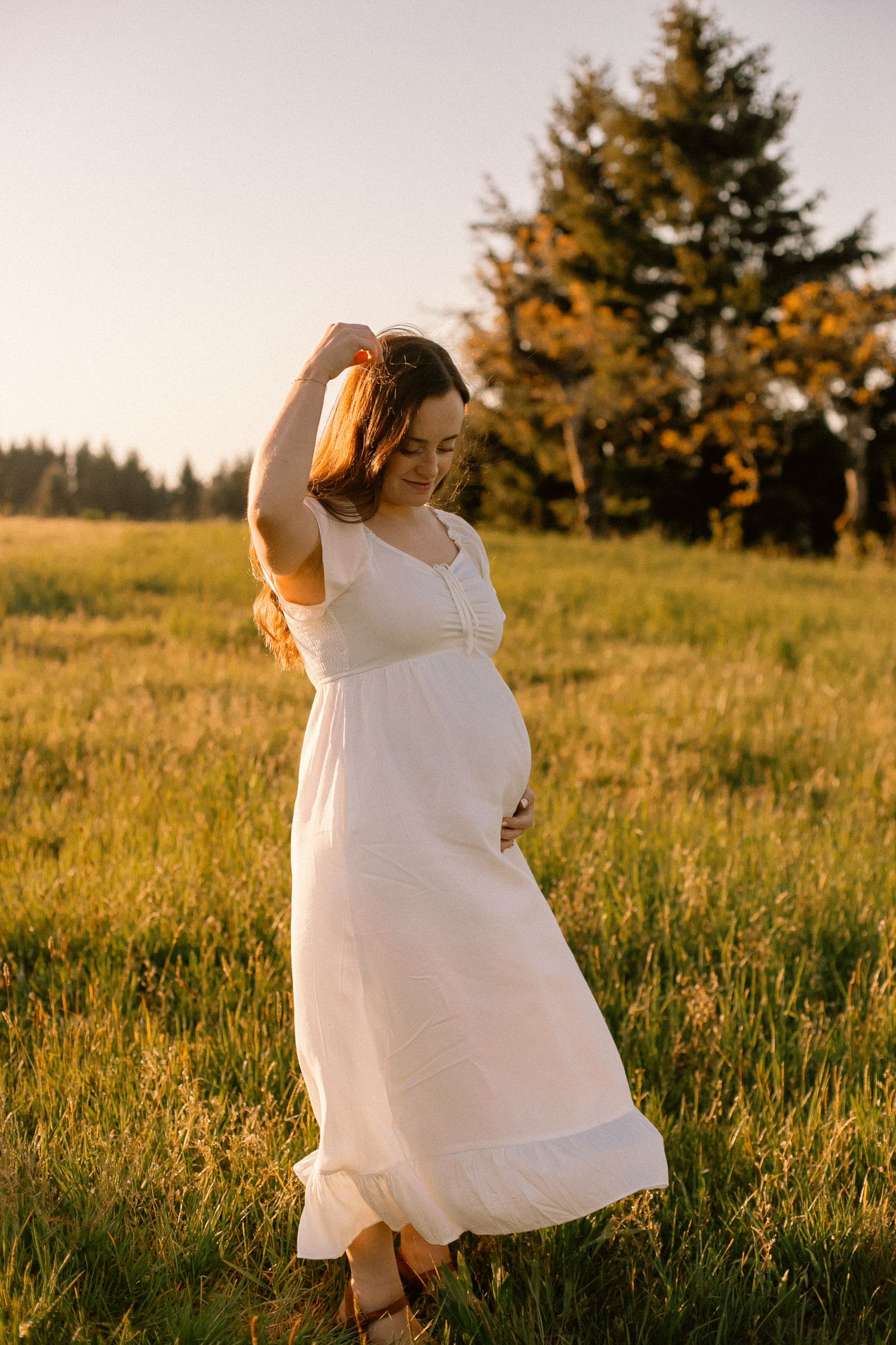 A young woman in a white dress twirls runs her fingers through her hair in a sunset lit field for a maternity photoshoot in Portland, Oregon