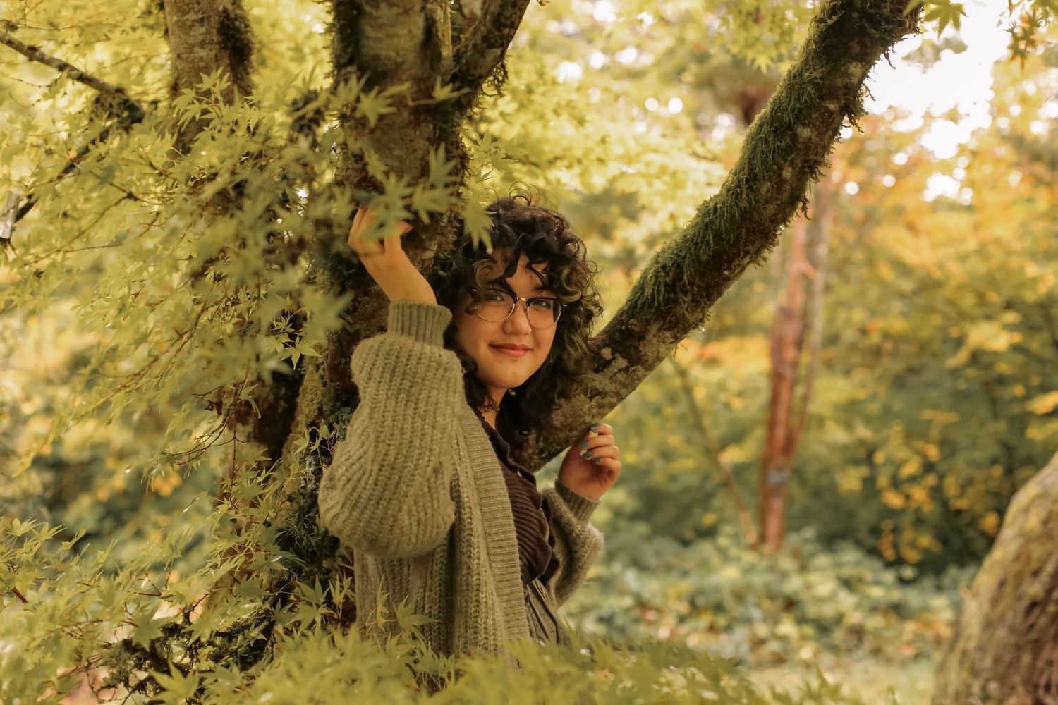 A young woman with dark, curly hair leaning against a tree in the forest with her hands on the branches.