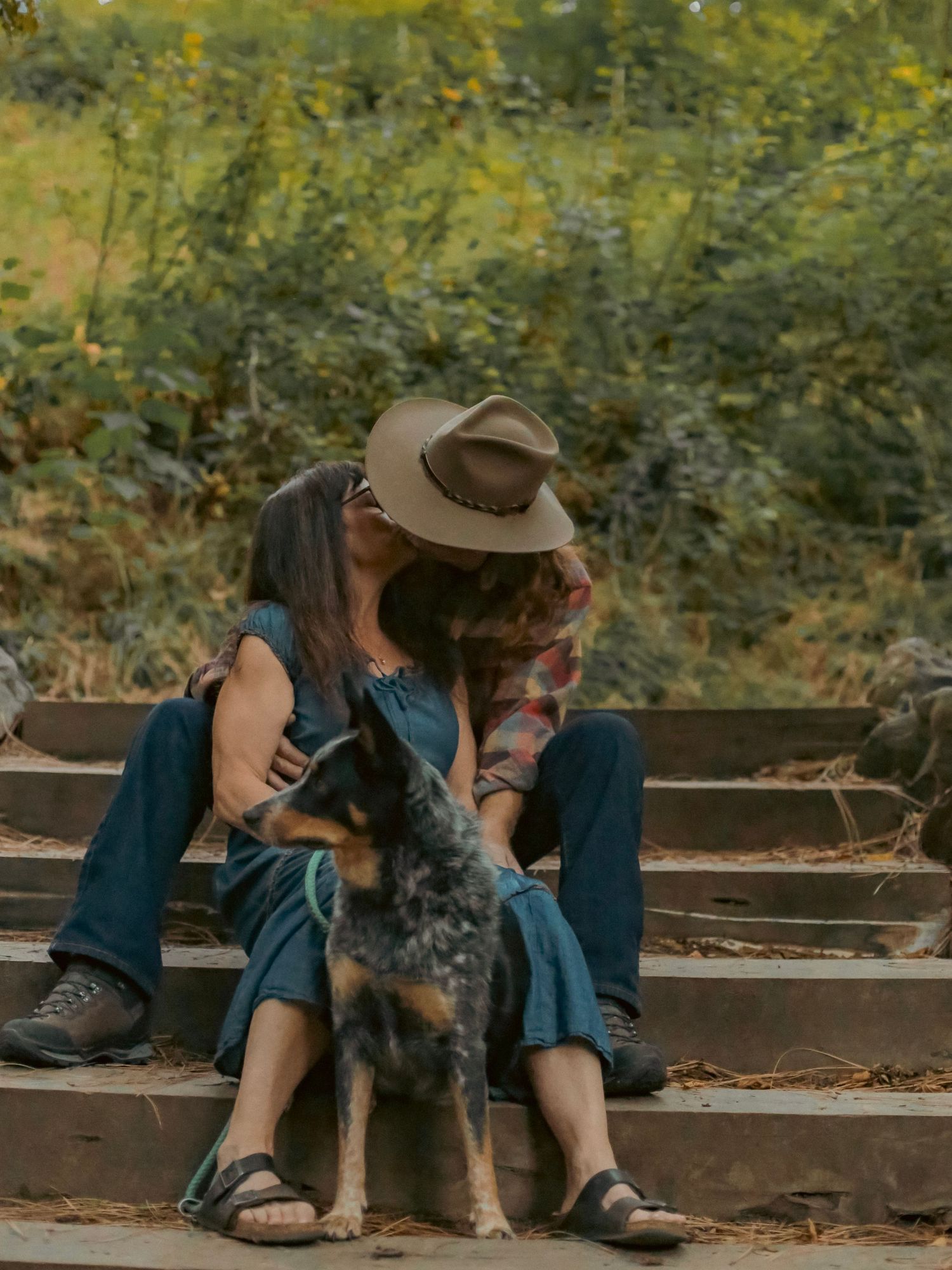 A man and a woman kissing and slightly hidden by the man's hat with their dog standing at their feet.