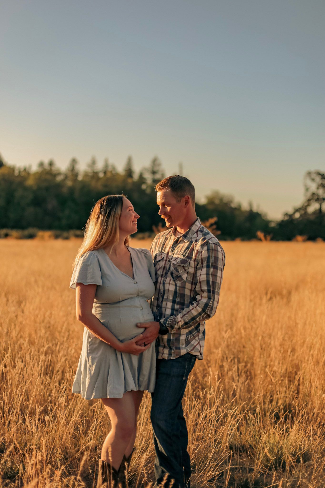 A man has his hand on his pregnant wife's belly in a sunny field while looking lovingly into each other's eyes