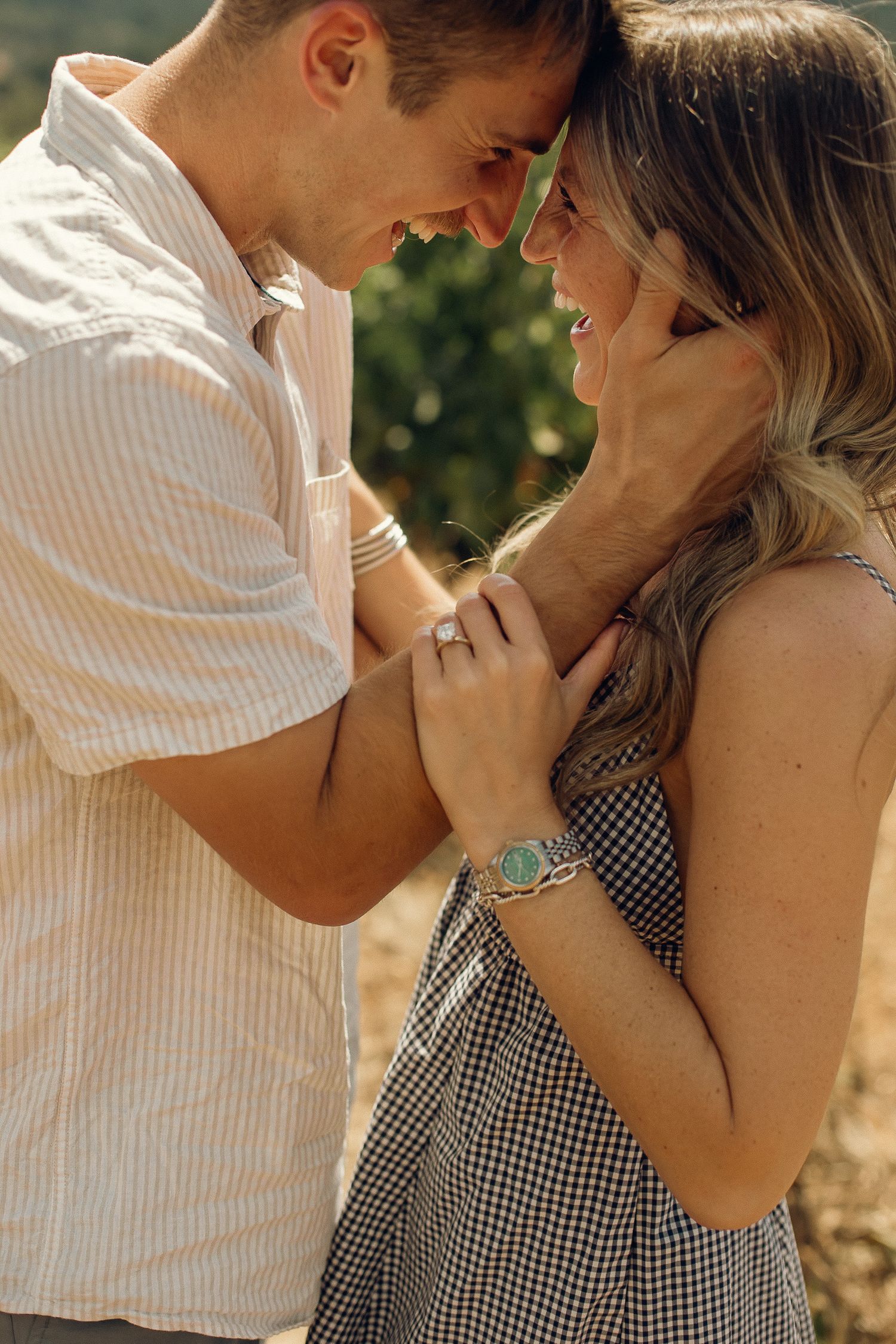 A newly engaged couple embraces with their foreheads together, laughing and smiling, in a sunny, warm vineyard in Oregon.