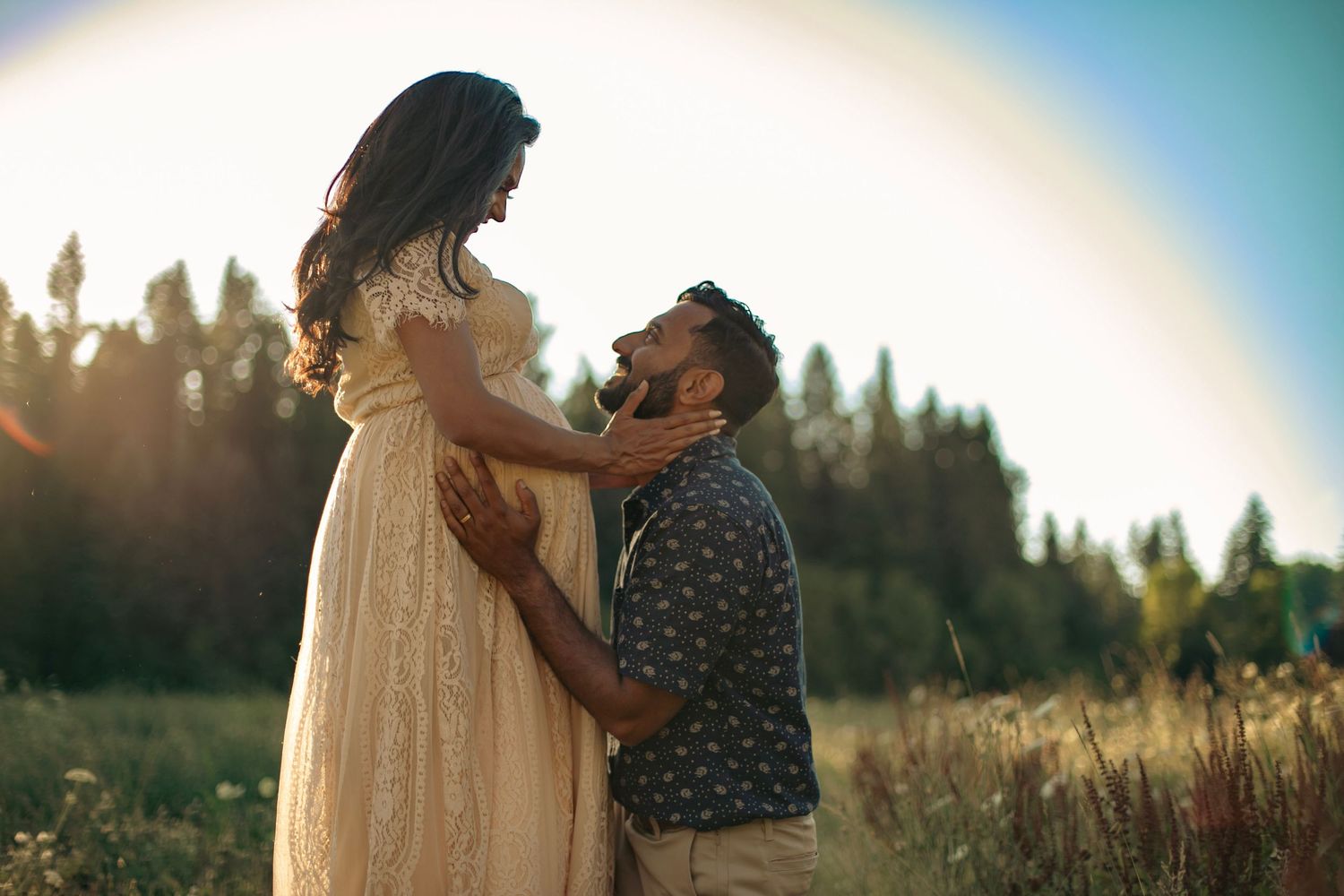 A pregnant woman and her husband looking at each other lovingly in a field with forest trees in the background.