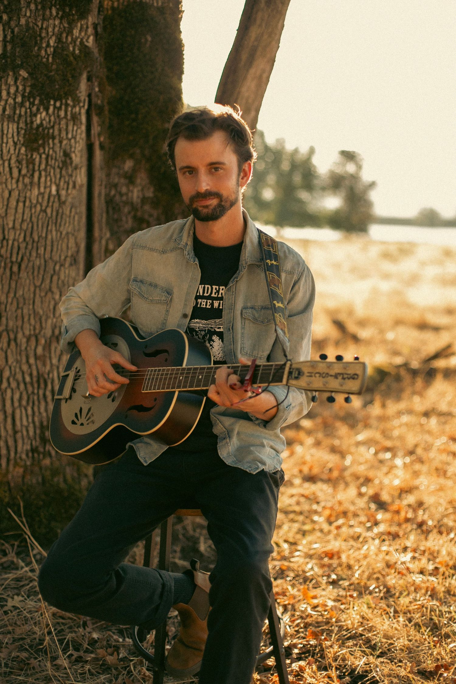 A man playing guitar and softly smiling at the camera while sitting on a stool in a grassy field.