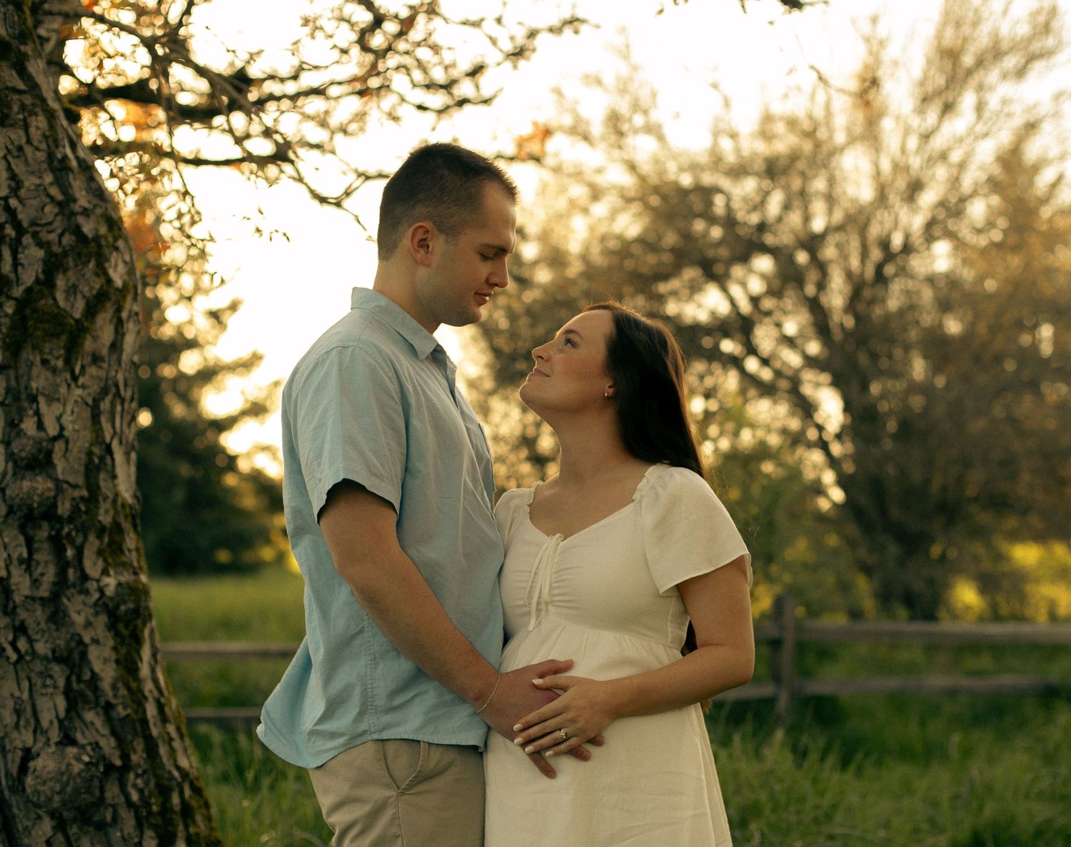 A couple poses and looks at each other in a field with tall trees for a cinematic, nostalgic, dreamy maternity photoshoot