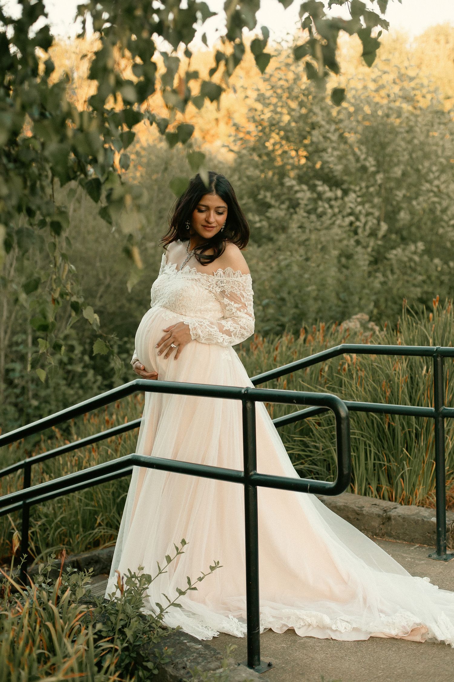 A woman poses holding her belly amongst green trees for a baby shower photoshoot in Southeast Portland, Oregon.