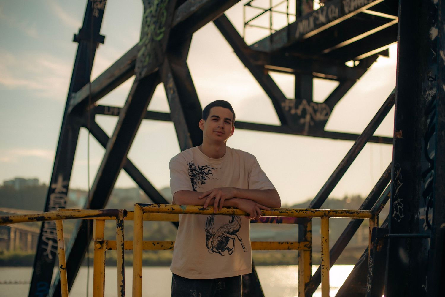 A young man posing in an industrial setting with a city river in the background.