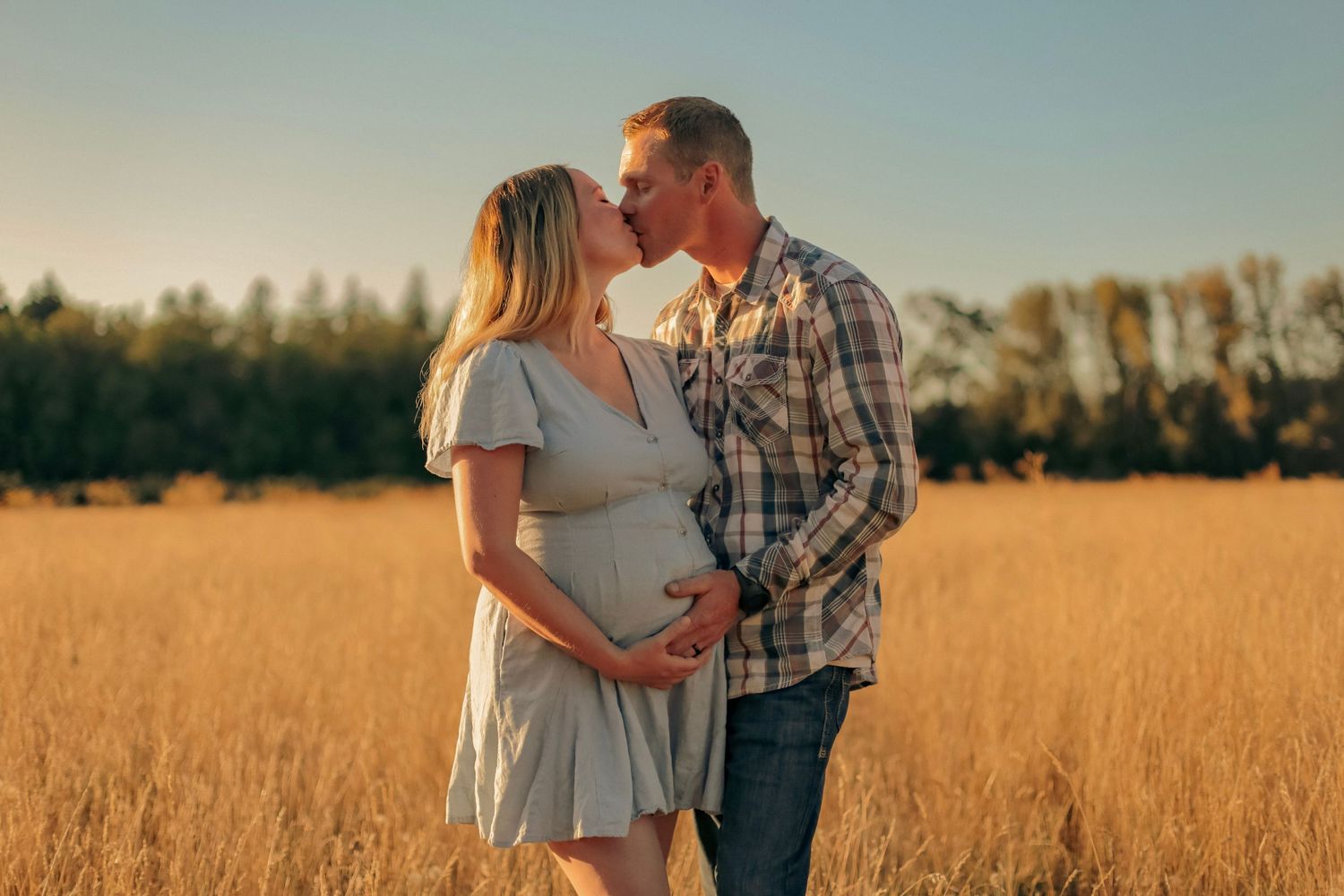 A woman and man kissing and holding her pregnant belly while standing in a golden wheat field.