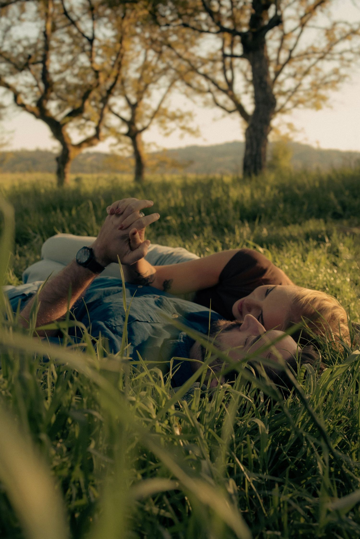 A couple laying in tall grass in a sunset-lit field holding hands