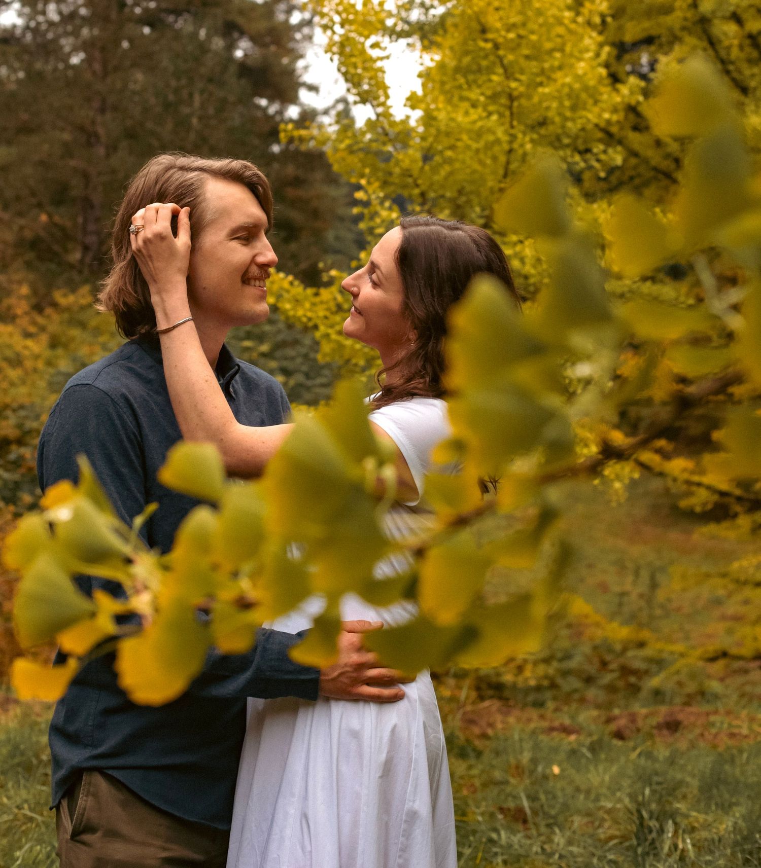 A woman in a white dress brushing the hair back from the face of her partner while looking at each other in between Ginkgo leaves.