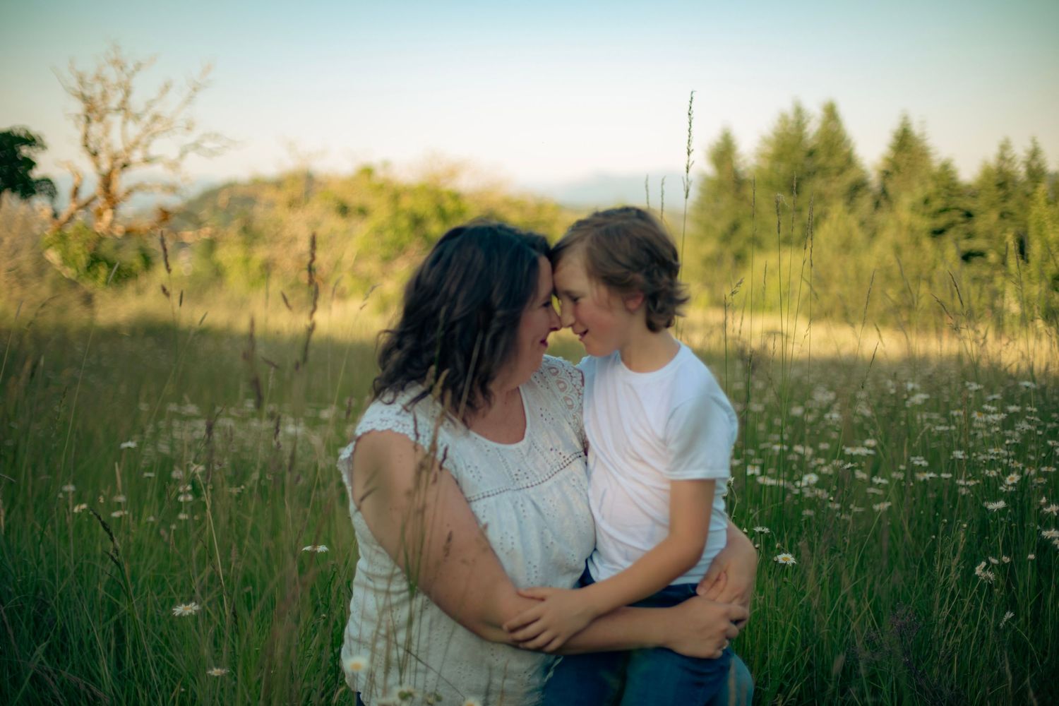 A mother and son sitting in a green field with white flowers looking at each other and touching foreheads.