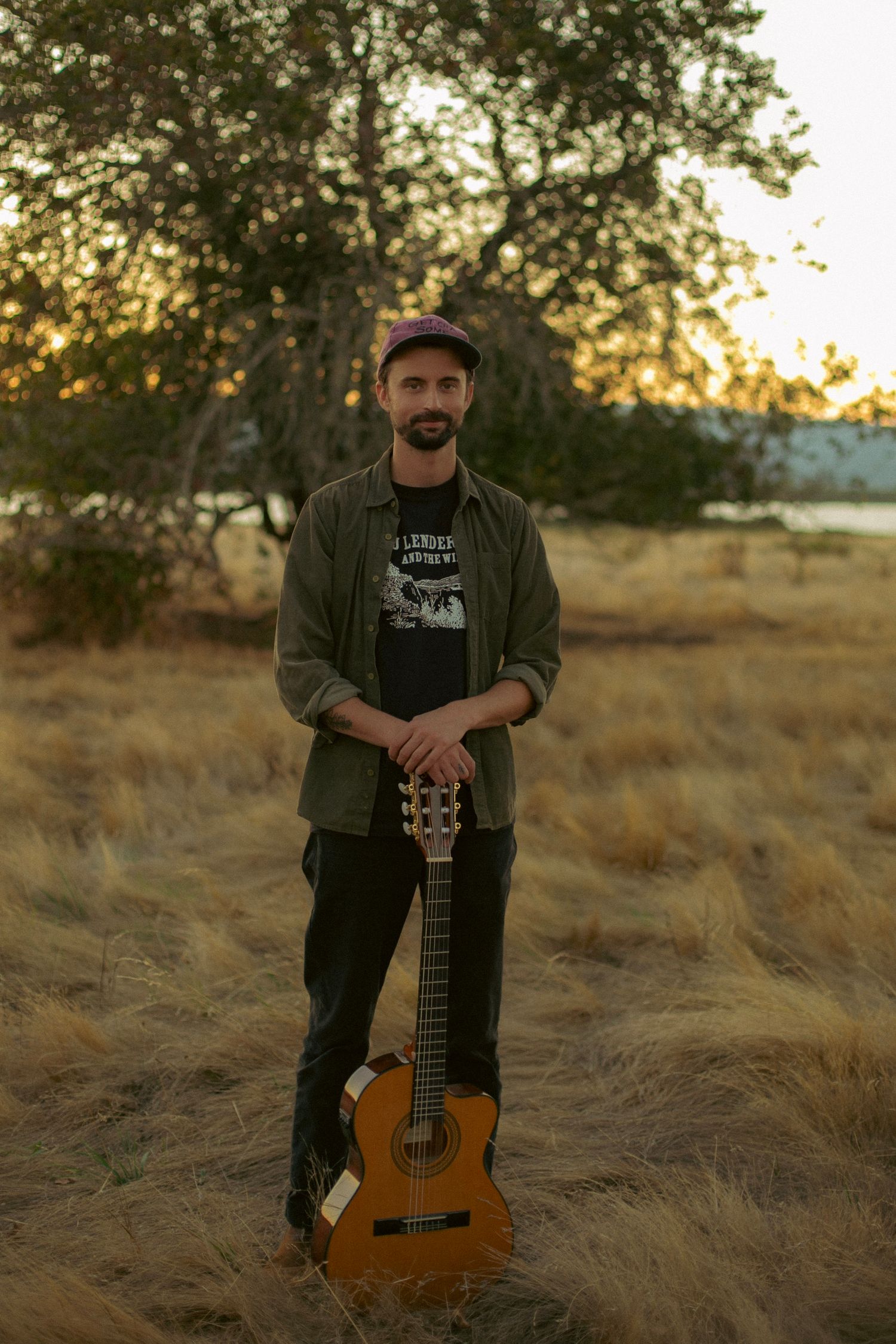 A man standing in a grassy field holding a guitar with the sun setting over a lake in the background.