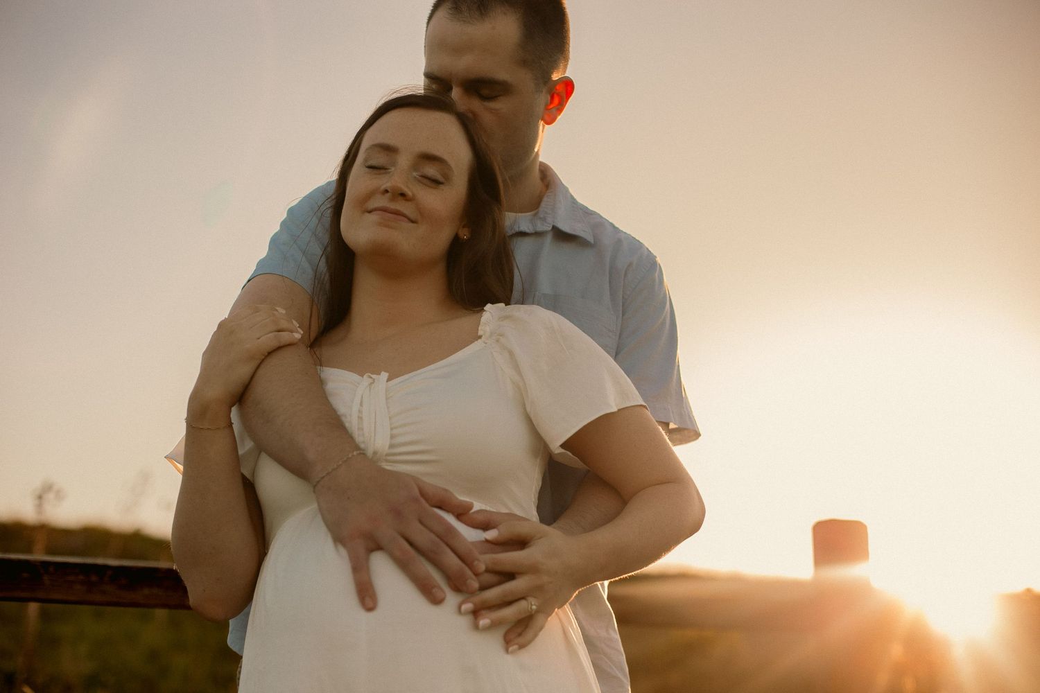 A couple embrace - the man has his hands around the woman from the back, holding her pregnant belly while they pose for a maternity photo