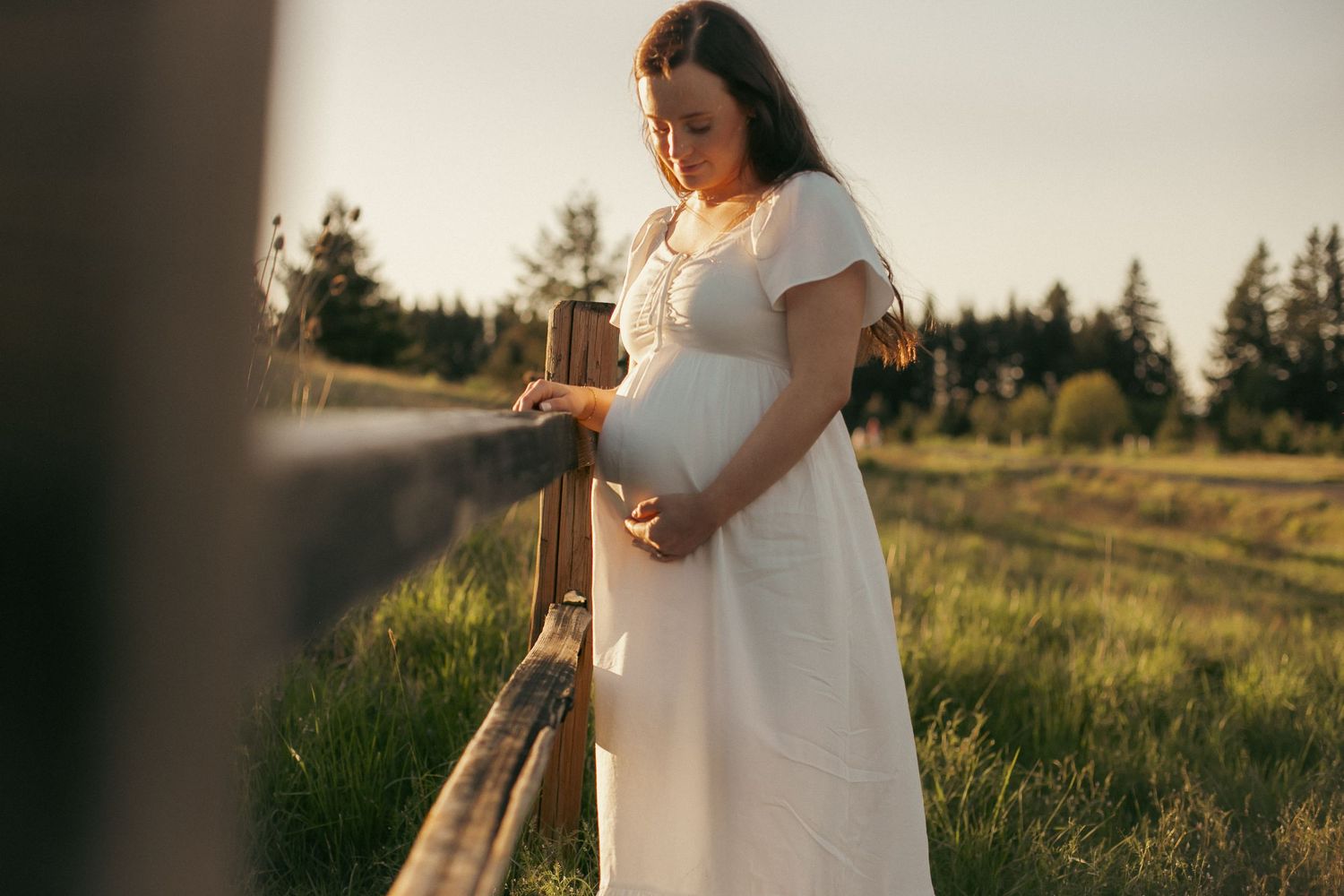 A woman in a white dress stands in a rural field leaning against a wooden fence while holding her pregnant belly for a maternity photoshoot