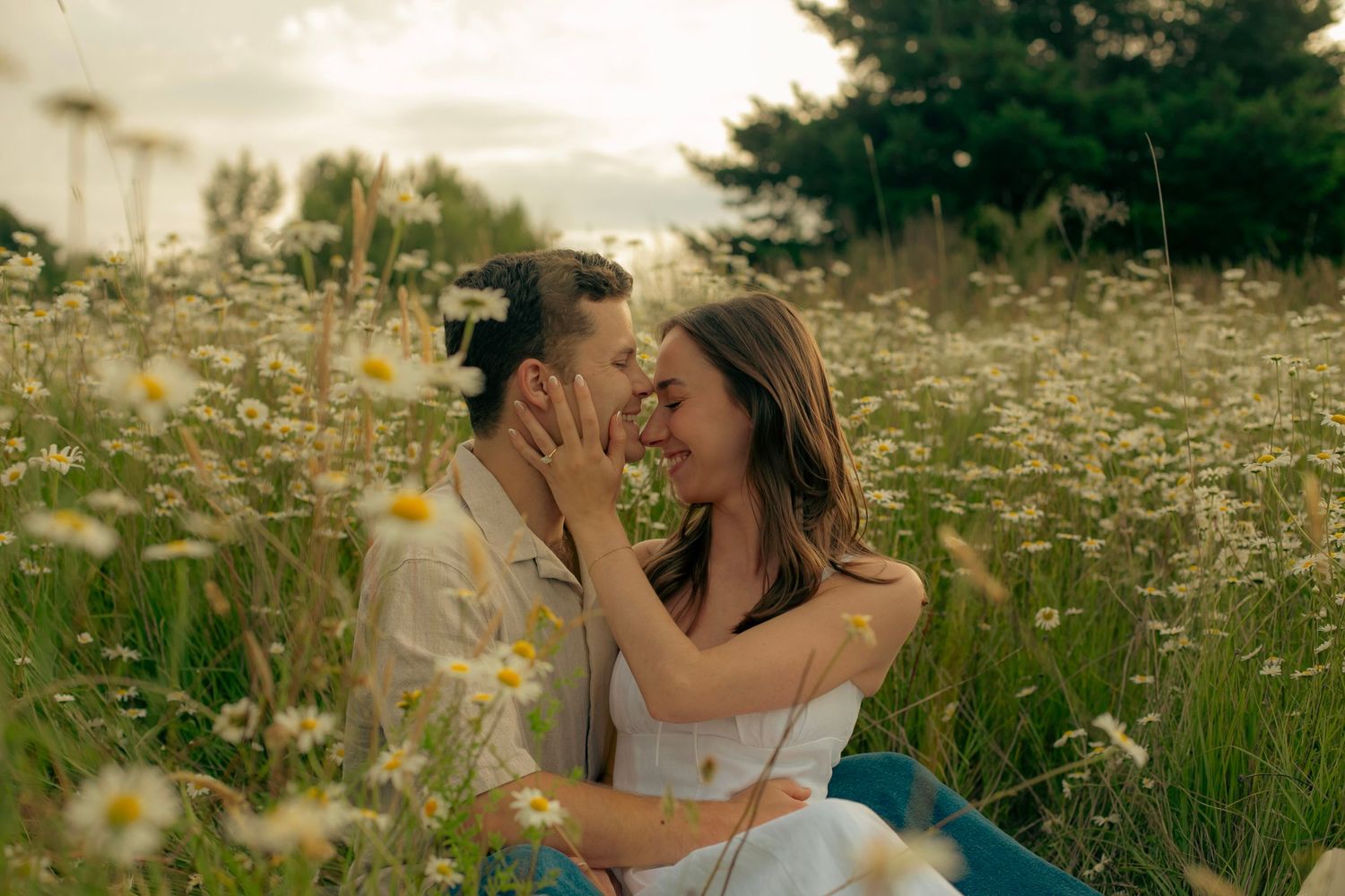 An engagement photoshoot shows a man and a woman sitting together in a field of wildflowers