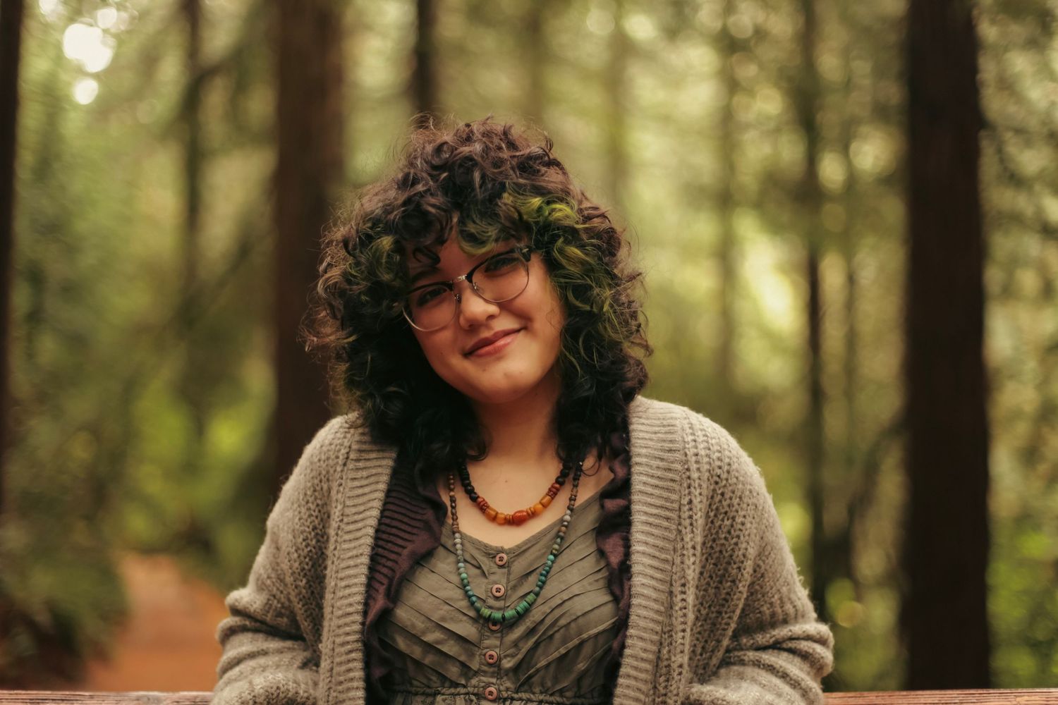 A young woman with dark curly hair and glasses smiling softly with redwood trees and greenery in the background.