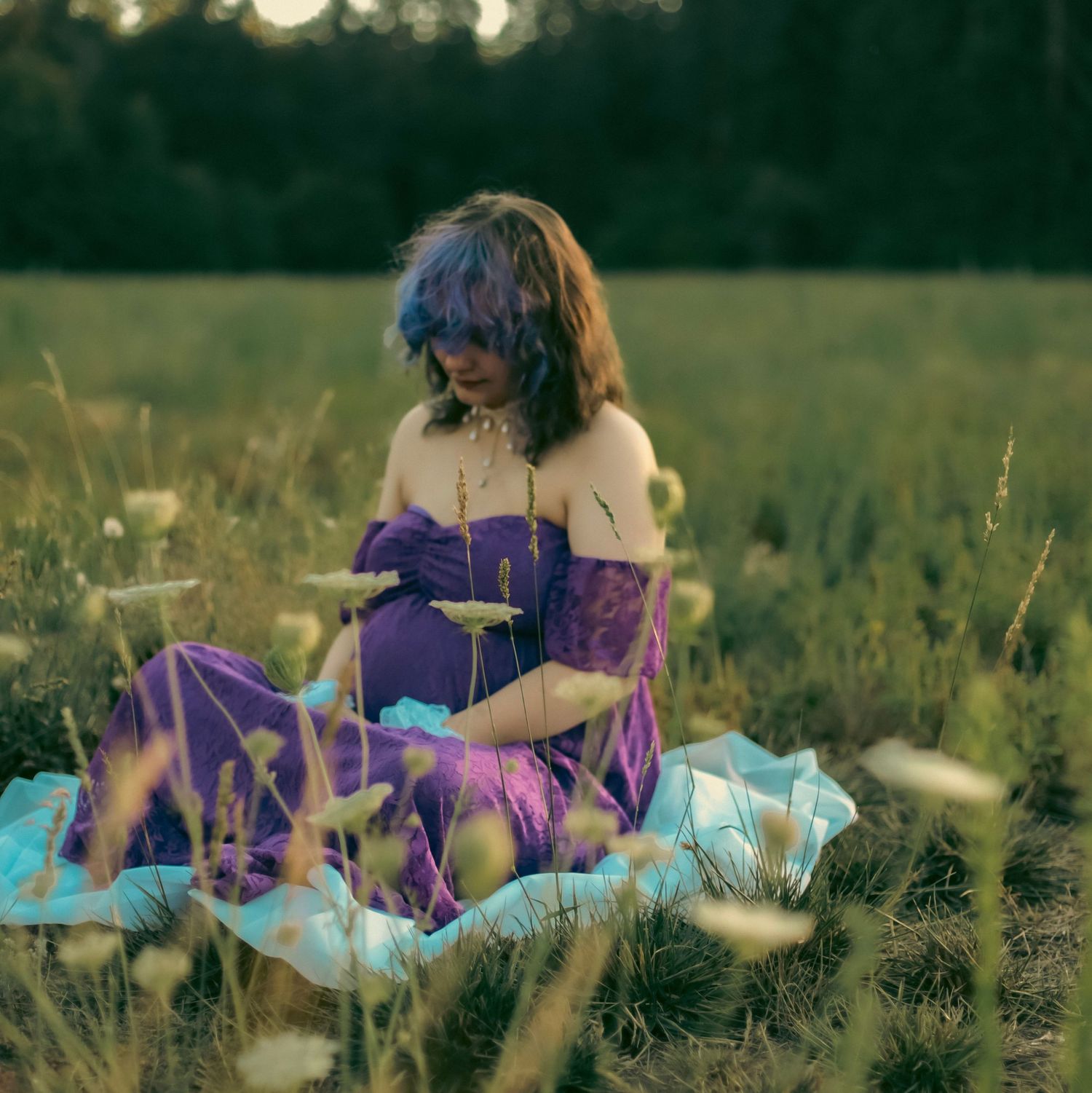 A maternity photo for a woman in a purple dress holding her belly and sitting amongst white flowers in a green grass field.