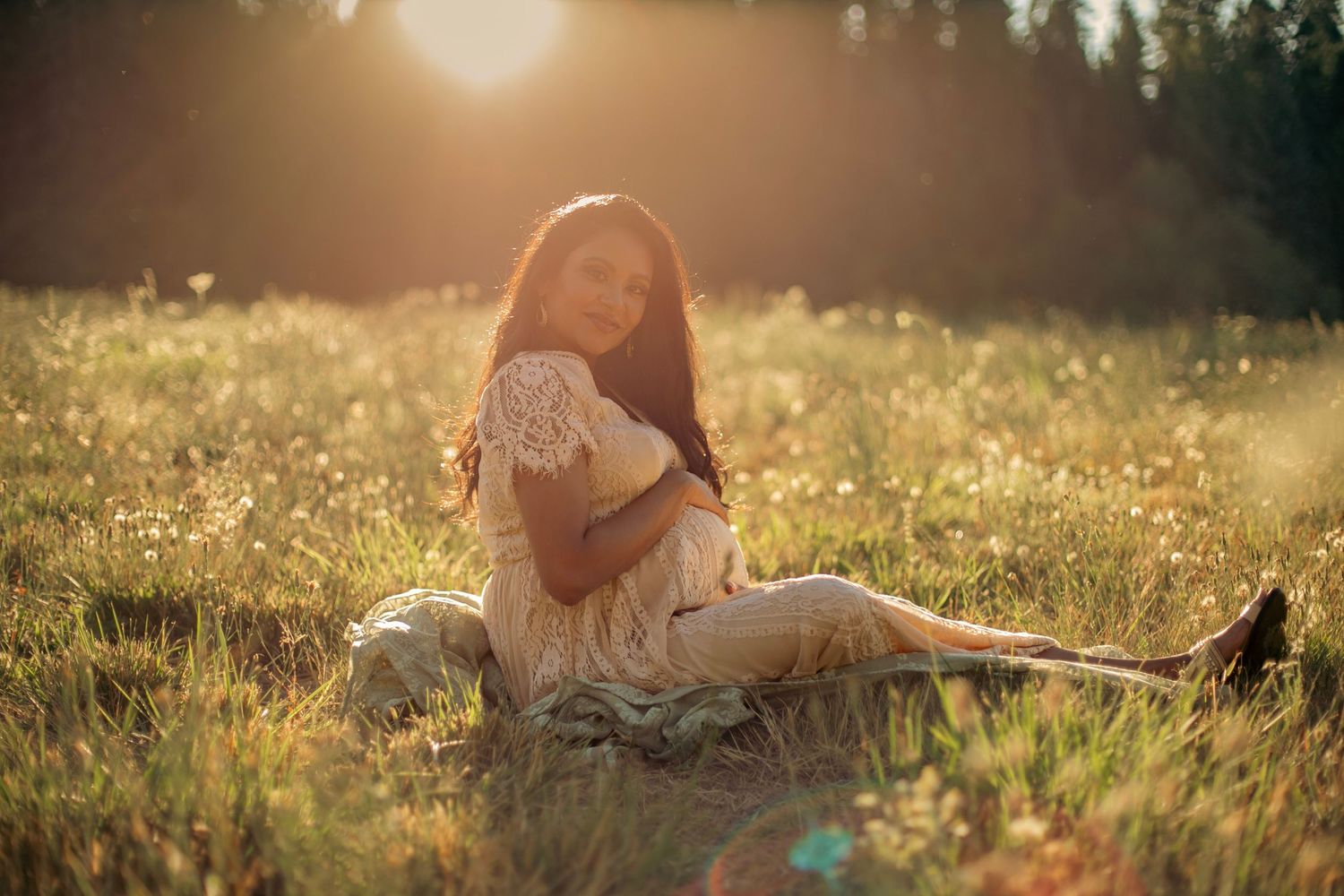 A maternity photo of a woman in a beautiful white dress holding her belly while sitting in a grassy field of wildflowers during sunset
