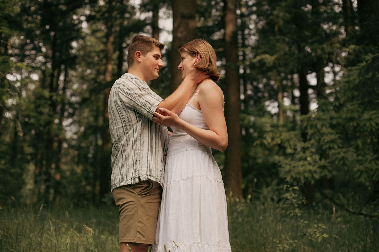 A couple looks into each other's eyes and poses with talk green trees in the background of a forest in Oregon for an engagement photoshoot