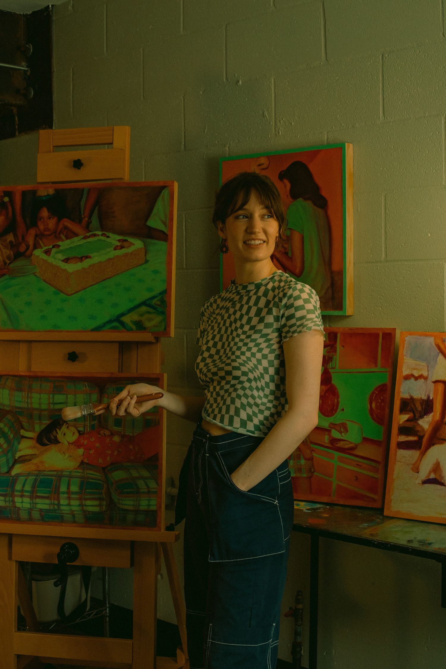 A woman poses in front of her paintings in her art studio holding a paintbrush and smiling for a solo portrait session in Portland, Oregon