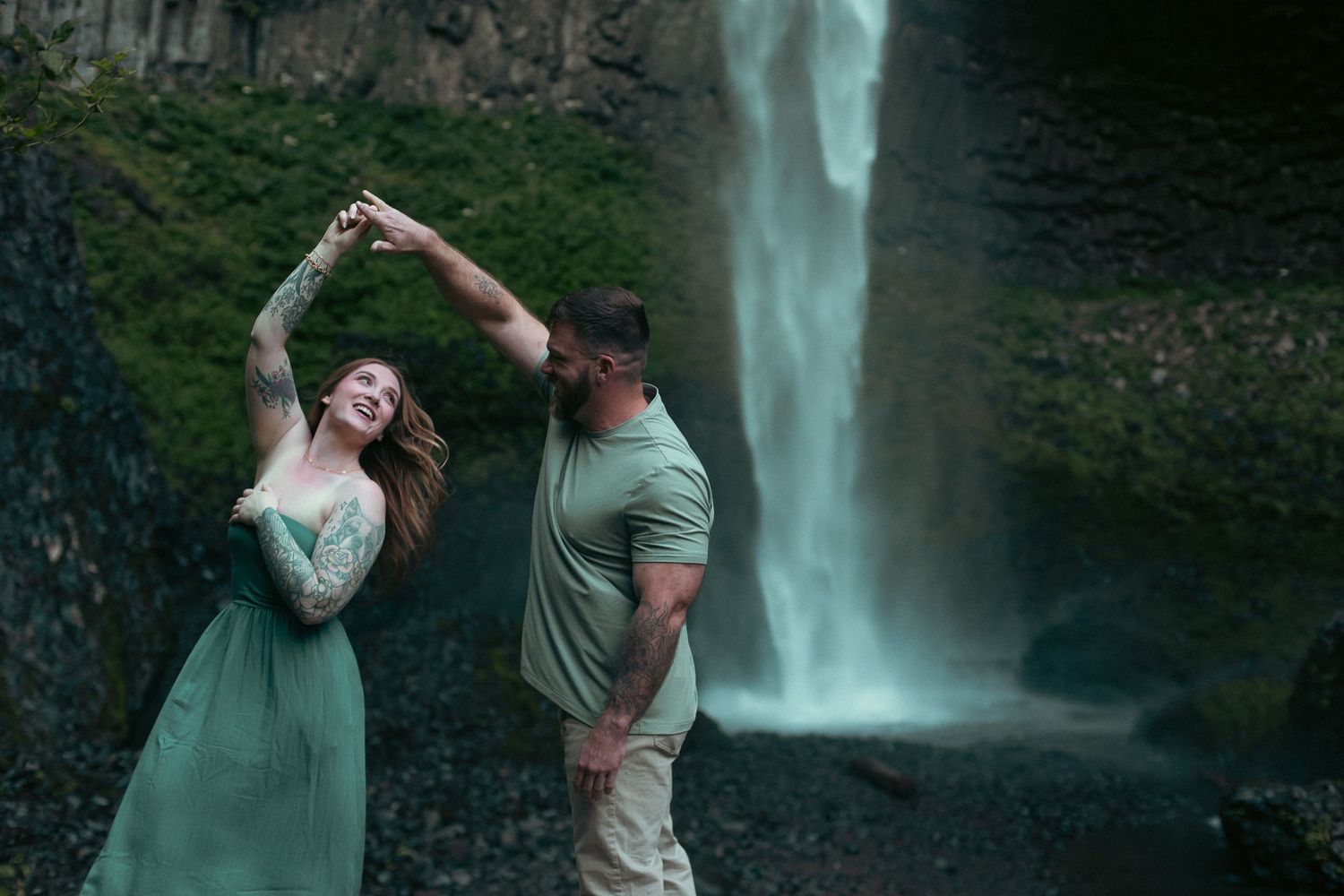 A couple wearing green holding hands and twirling in front of a rushing waterfall for a moody engagement photoshoot in Oregon