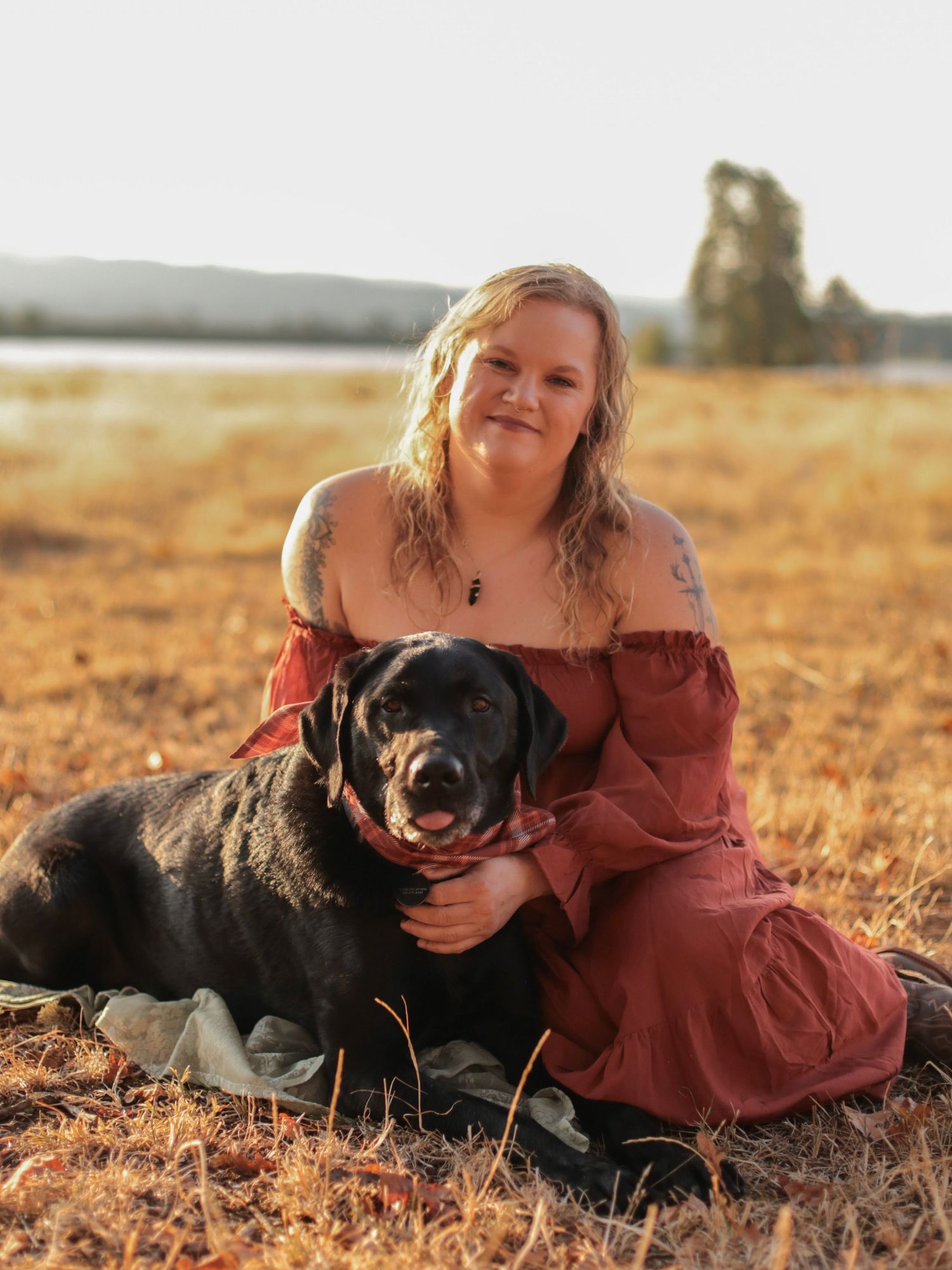 A woman in a red dress holding her black dog while sitting out in the country with the sunset in the background.