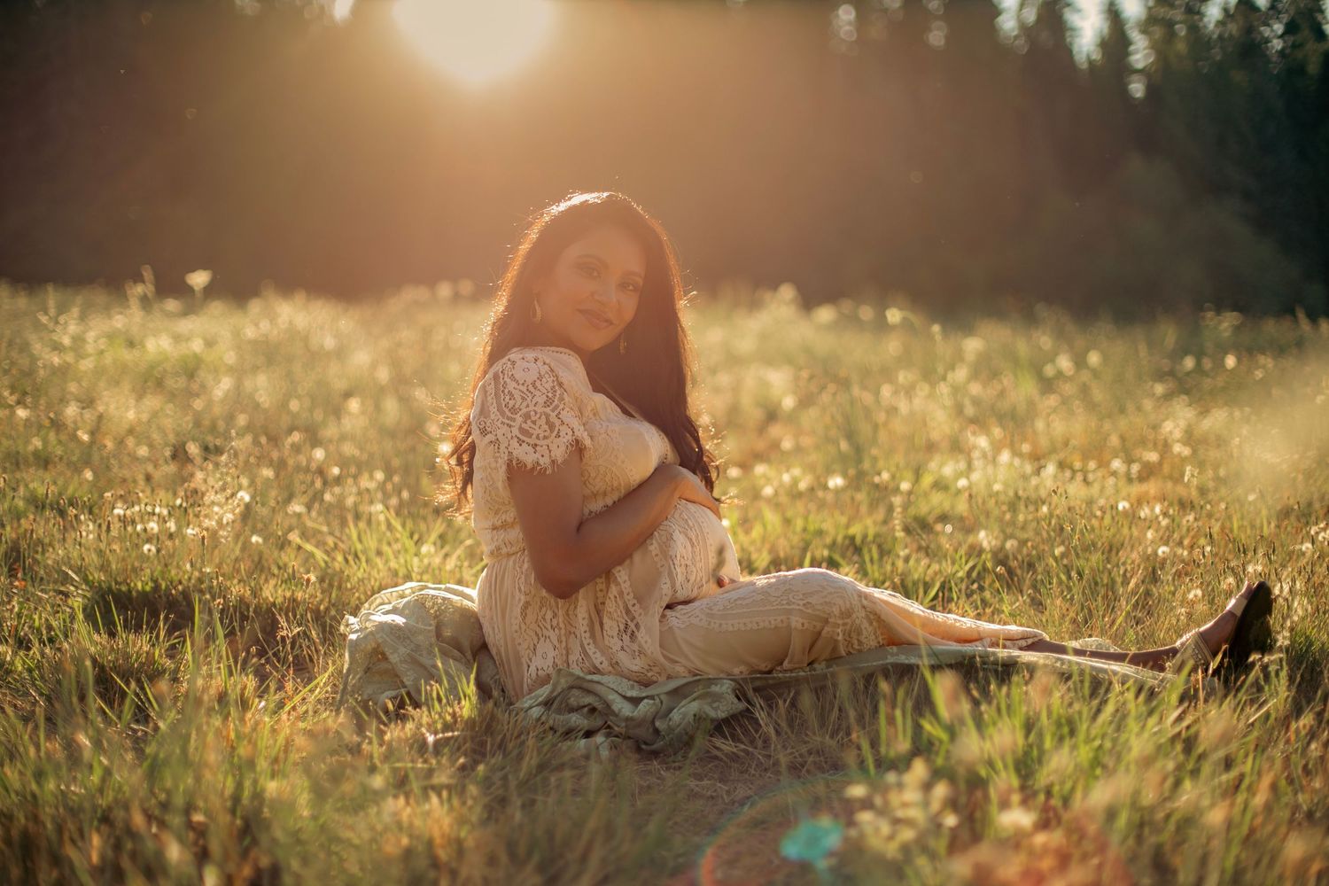 A maternity photo of a woman in a lacey, white dress holding her belly while sitting in a field of white flowers with the sun setting.