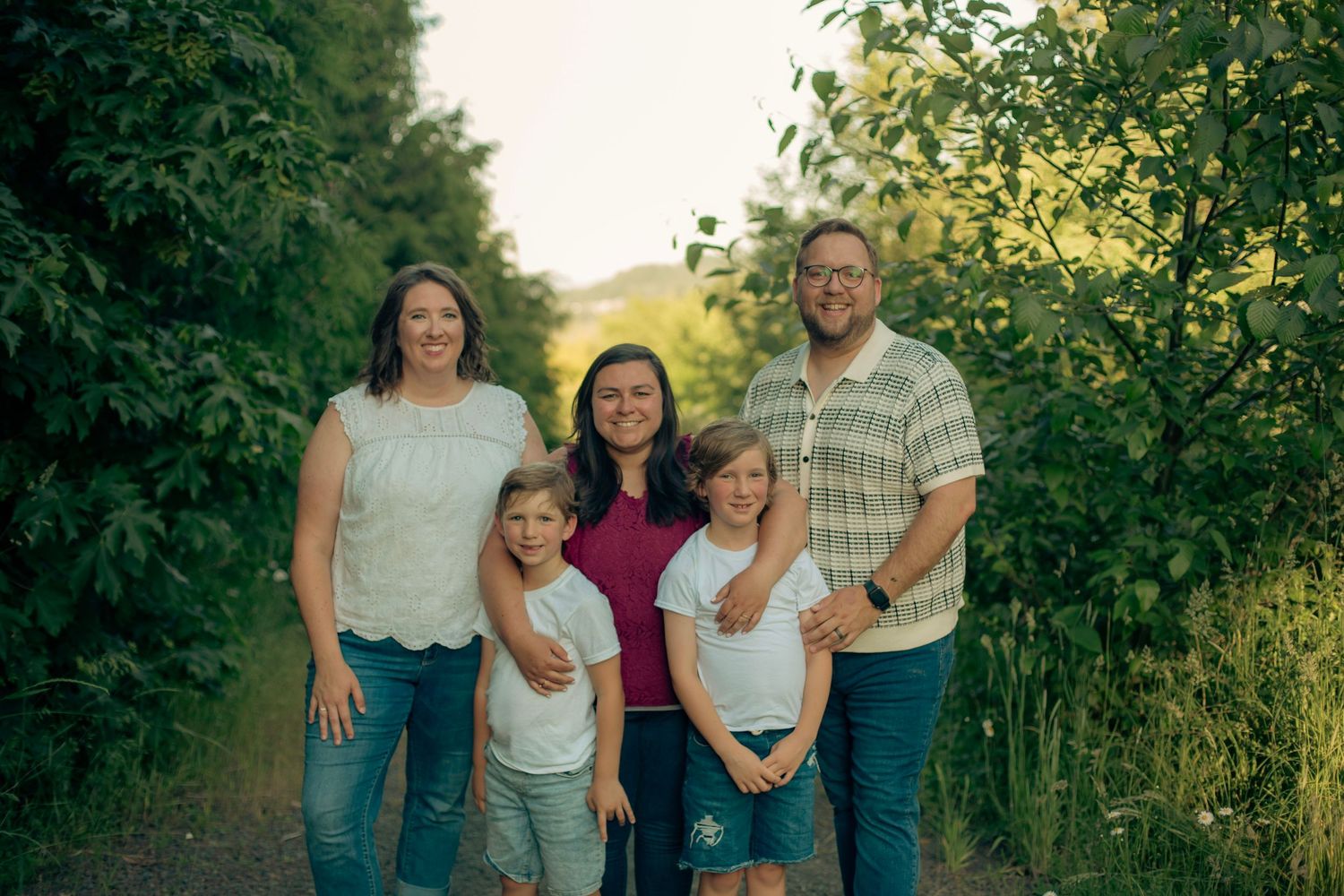 A family of five smiling and standing close to each other with green trees, blue sky and a mountain in the background.