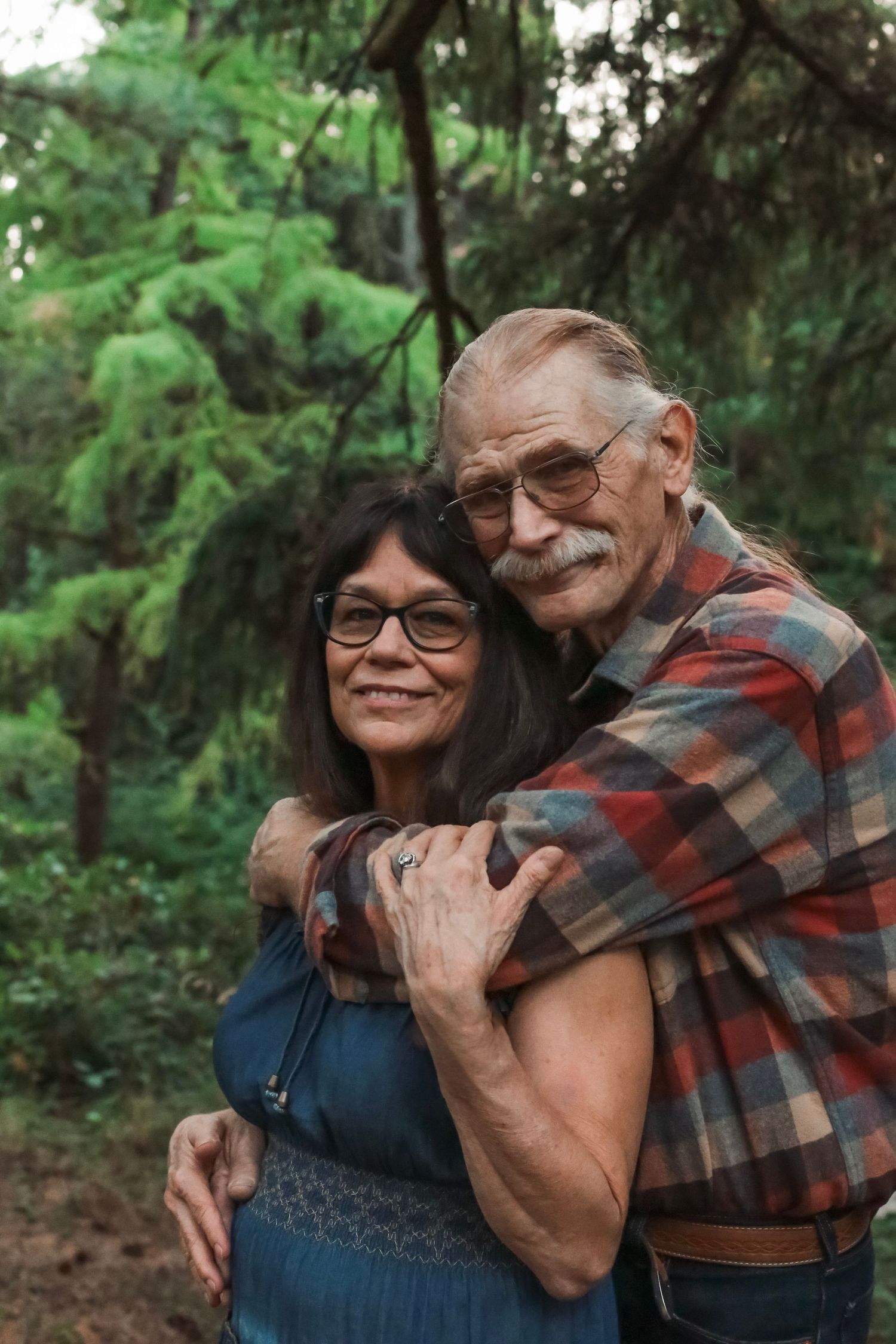An older couple with their arms around each other smiling at the camera with green trees in the background.