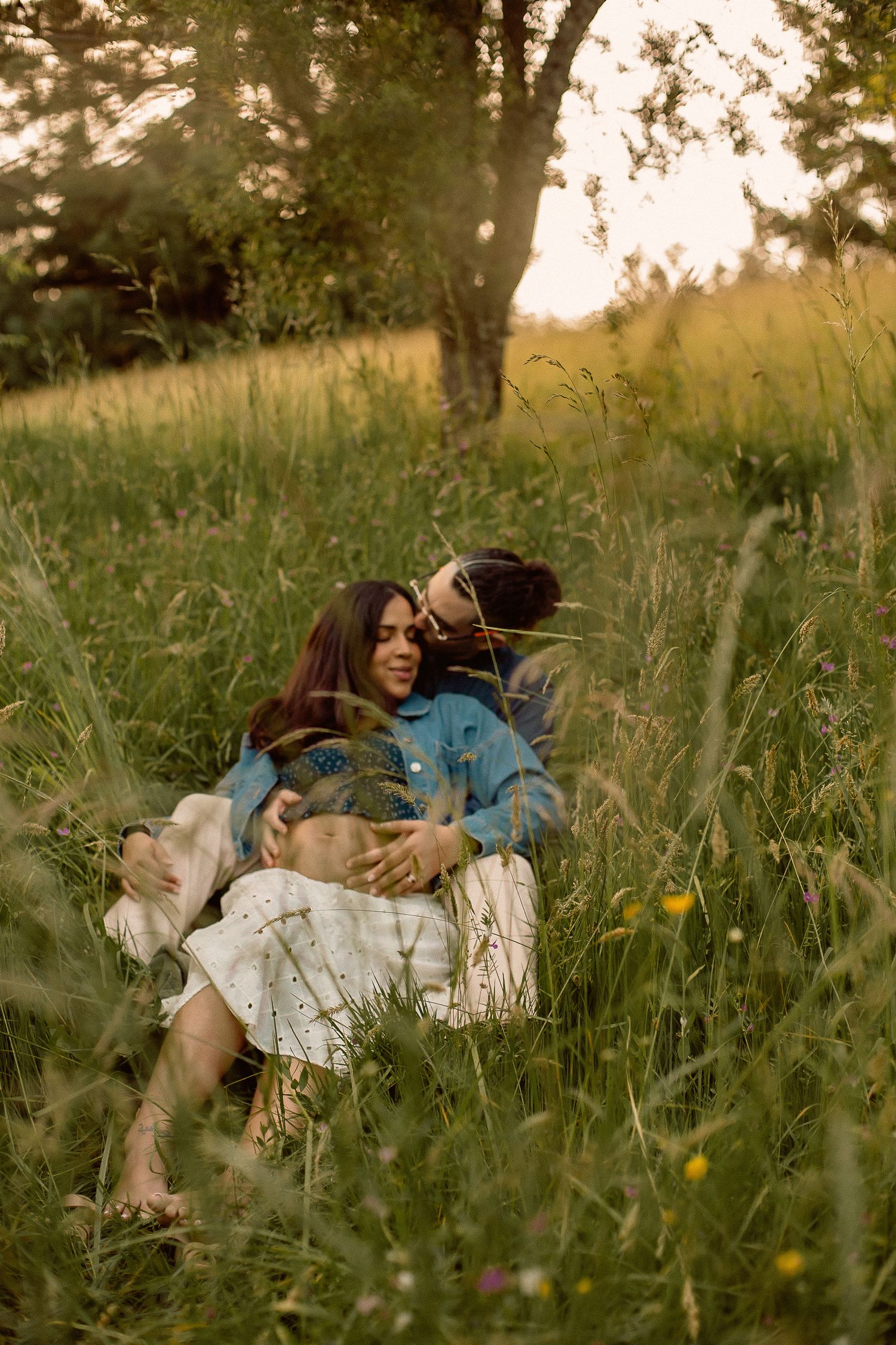 A couple sits in a tall grass field with the woman leaning into the man's lap for a maternity photoshoot