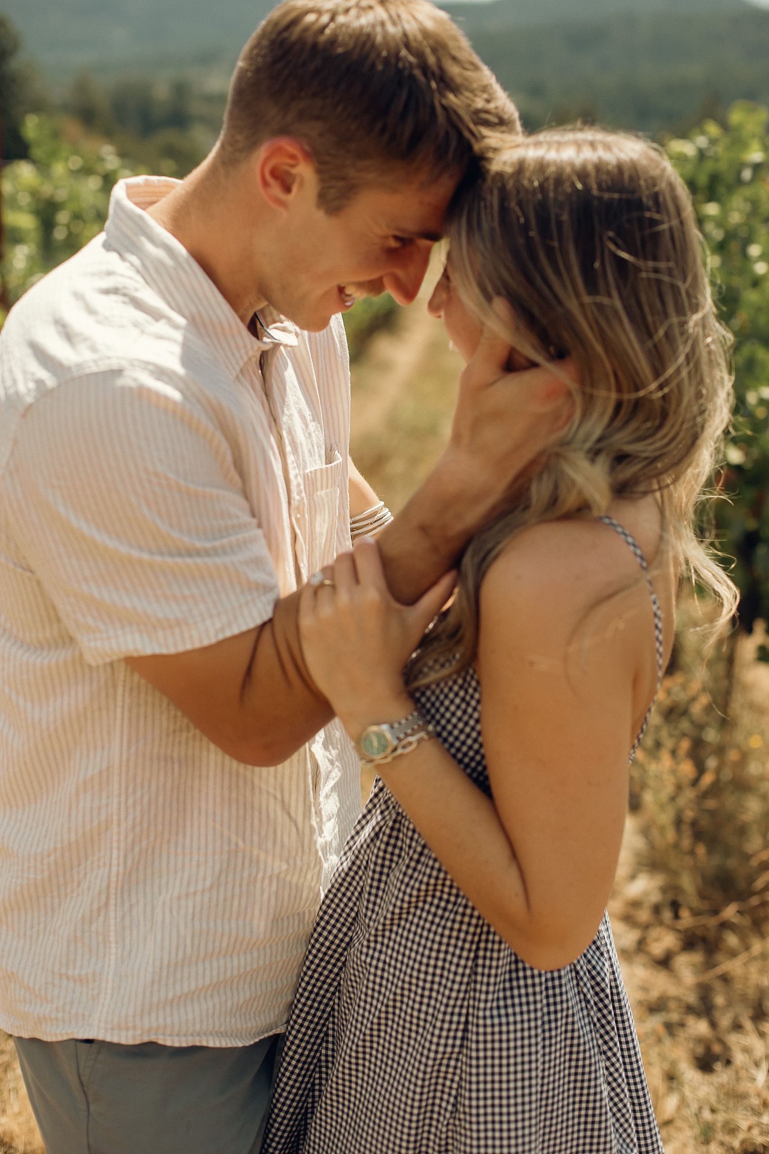 A newly engaged couple embraces with their foreheads together, laughing and smiling, in a sunny, warm vineyard in Oregon.