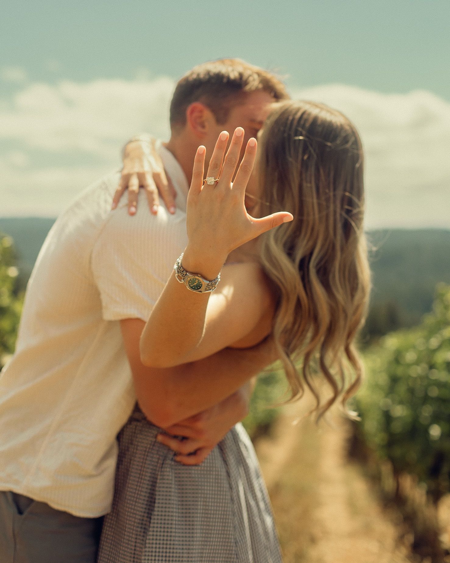 A newly engaged couple embrace and kiss while the woman holds her hand out in front of them, showing her new engagement ring.