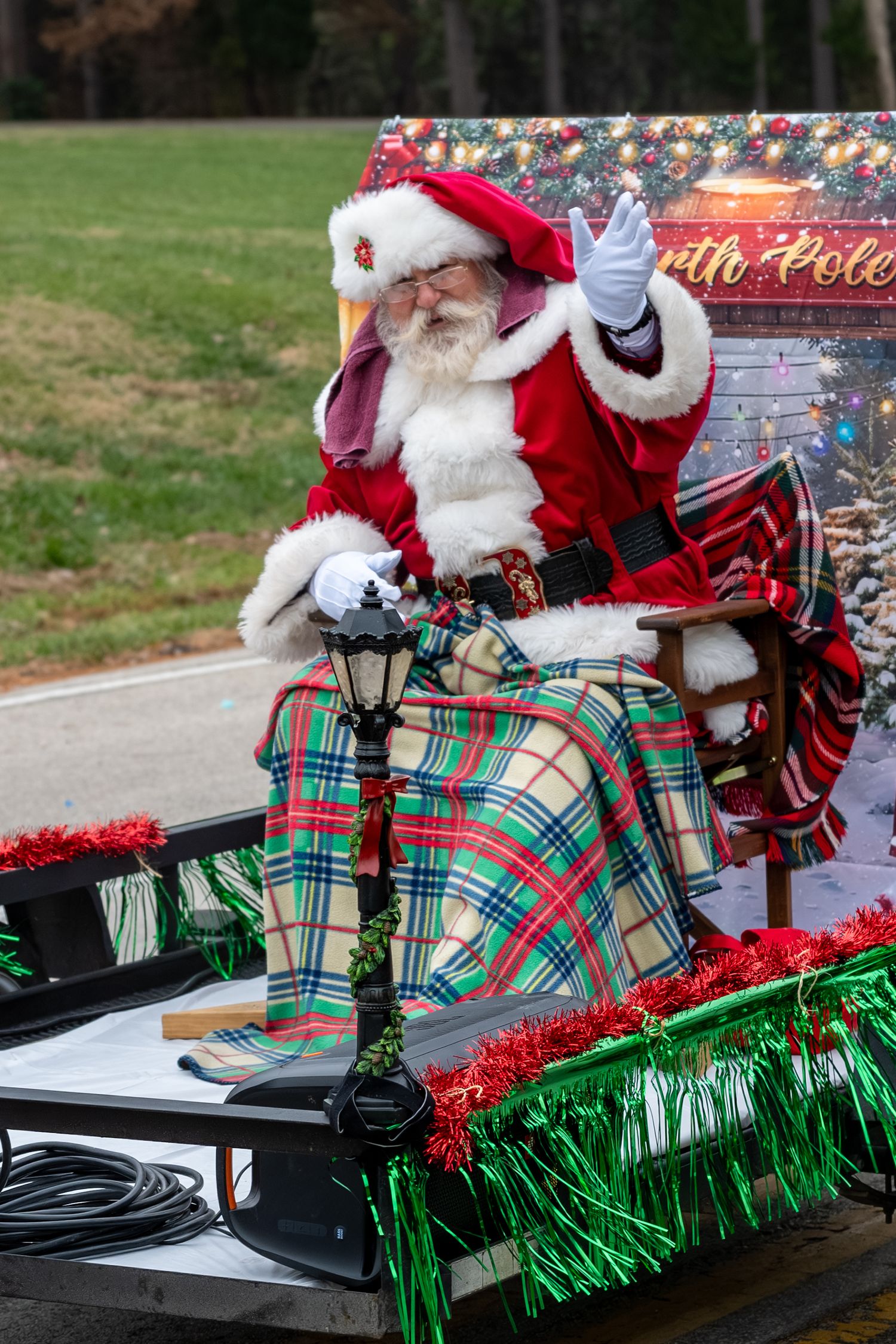 Santa Claus waves from a festive parade float decorated with green tinsel and a plaid blanket during a holiday parade.