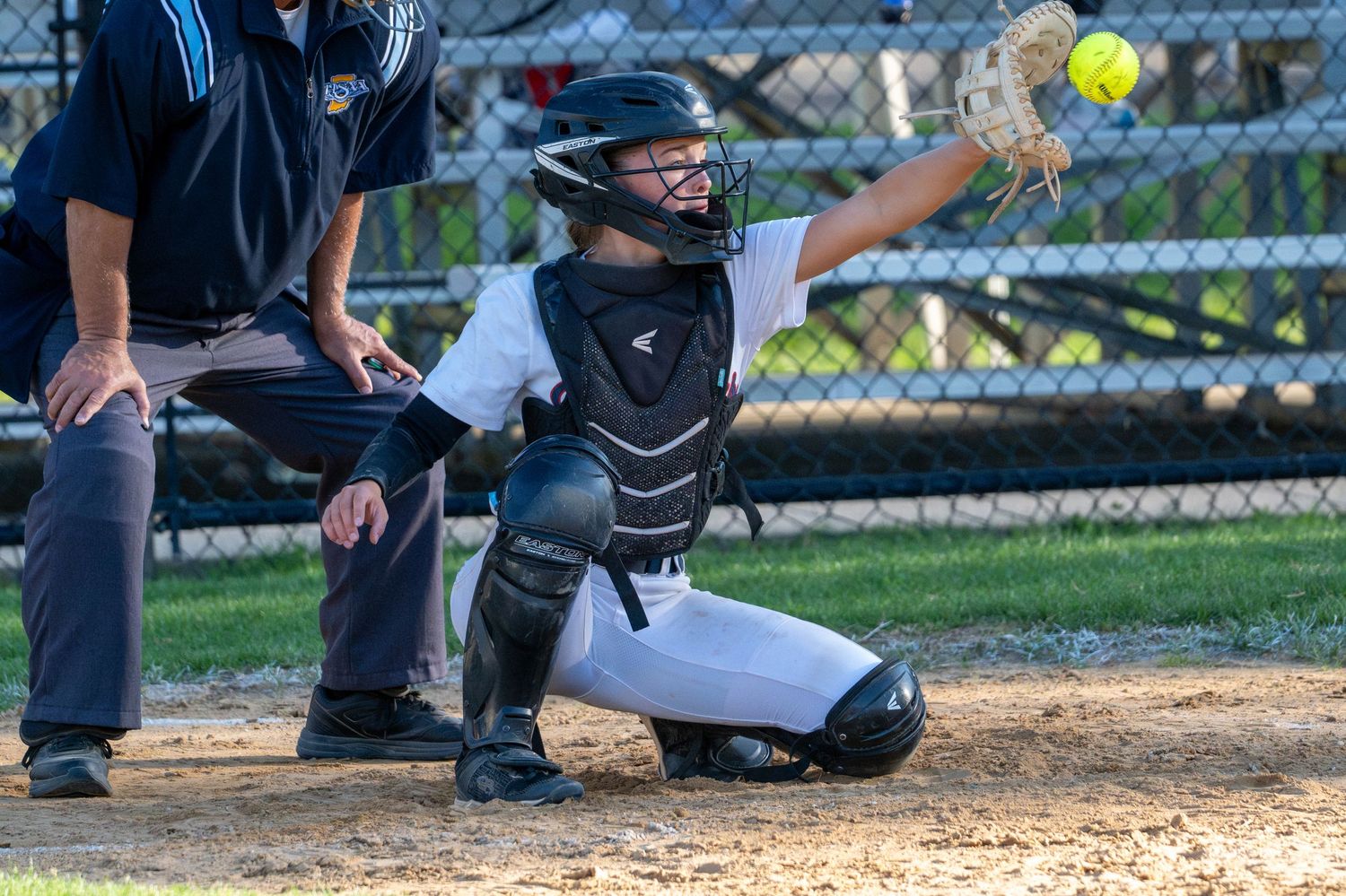 Youth softball catcher in full gear crouching at home plate, throwing ball during a game.