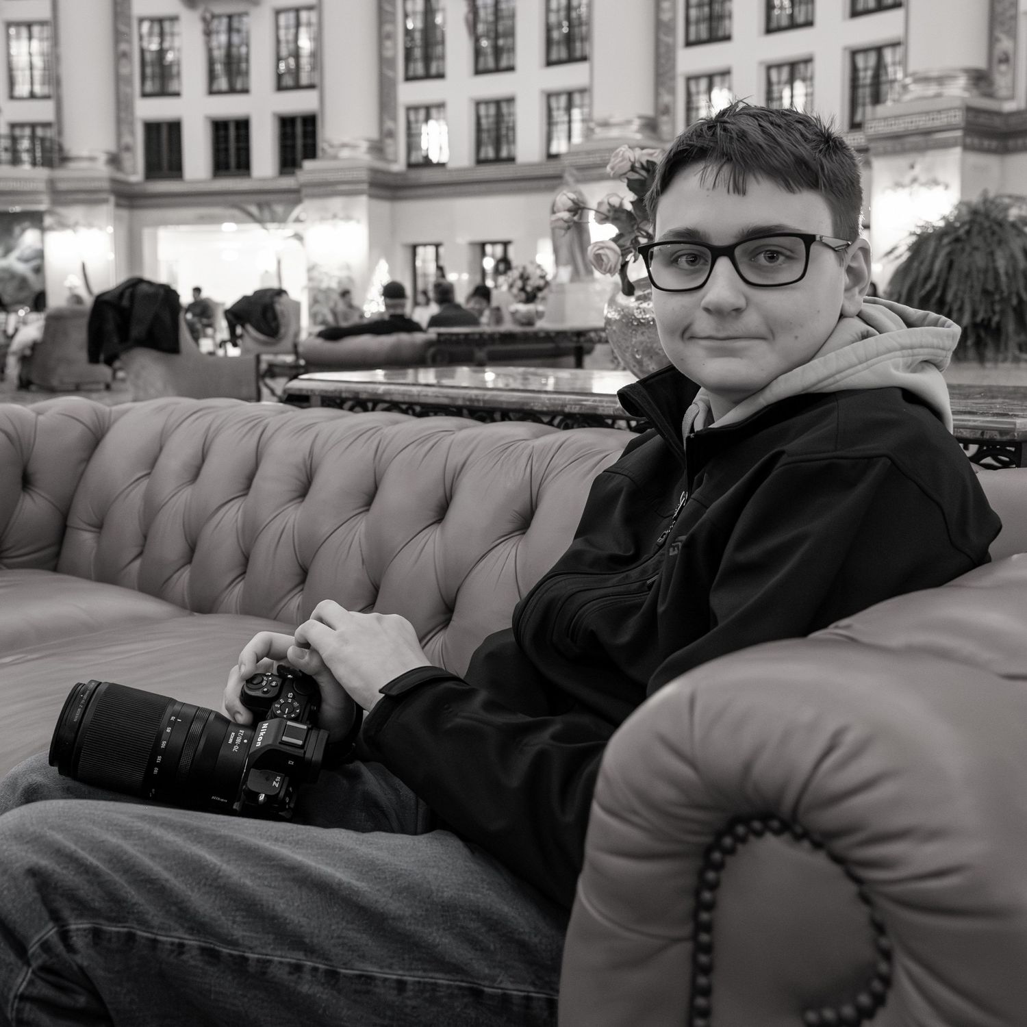 Young photographer with glasses sits on tufted sofa holding camera in black and white urban setting.