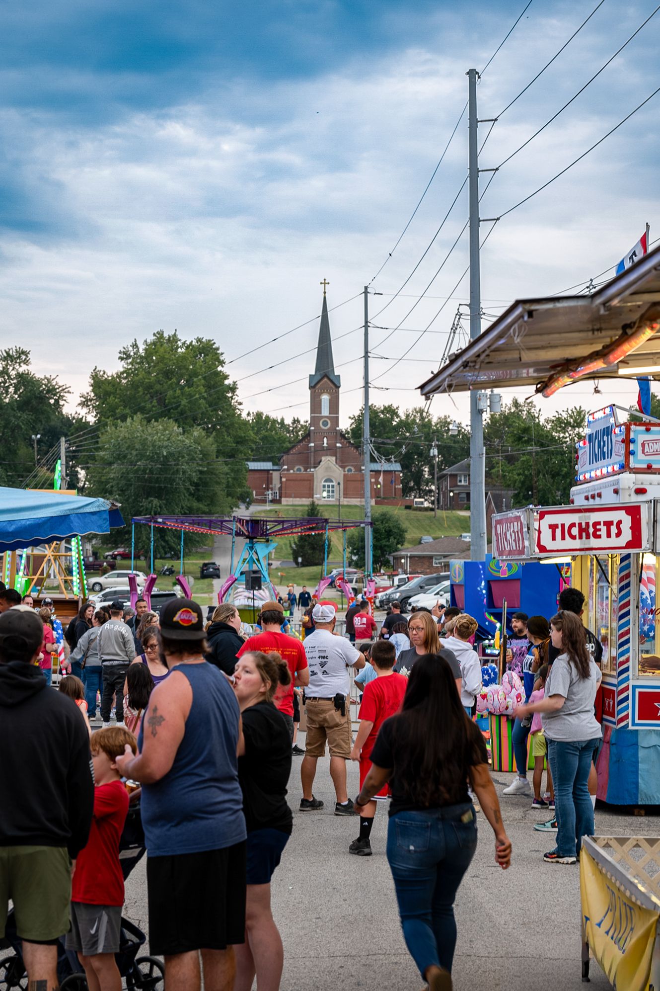 Crowds enjoy a lively outdoor carnival with ticket booths, rides, and a church steeple visible in the background.