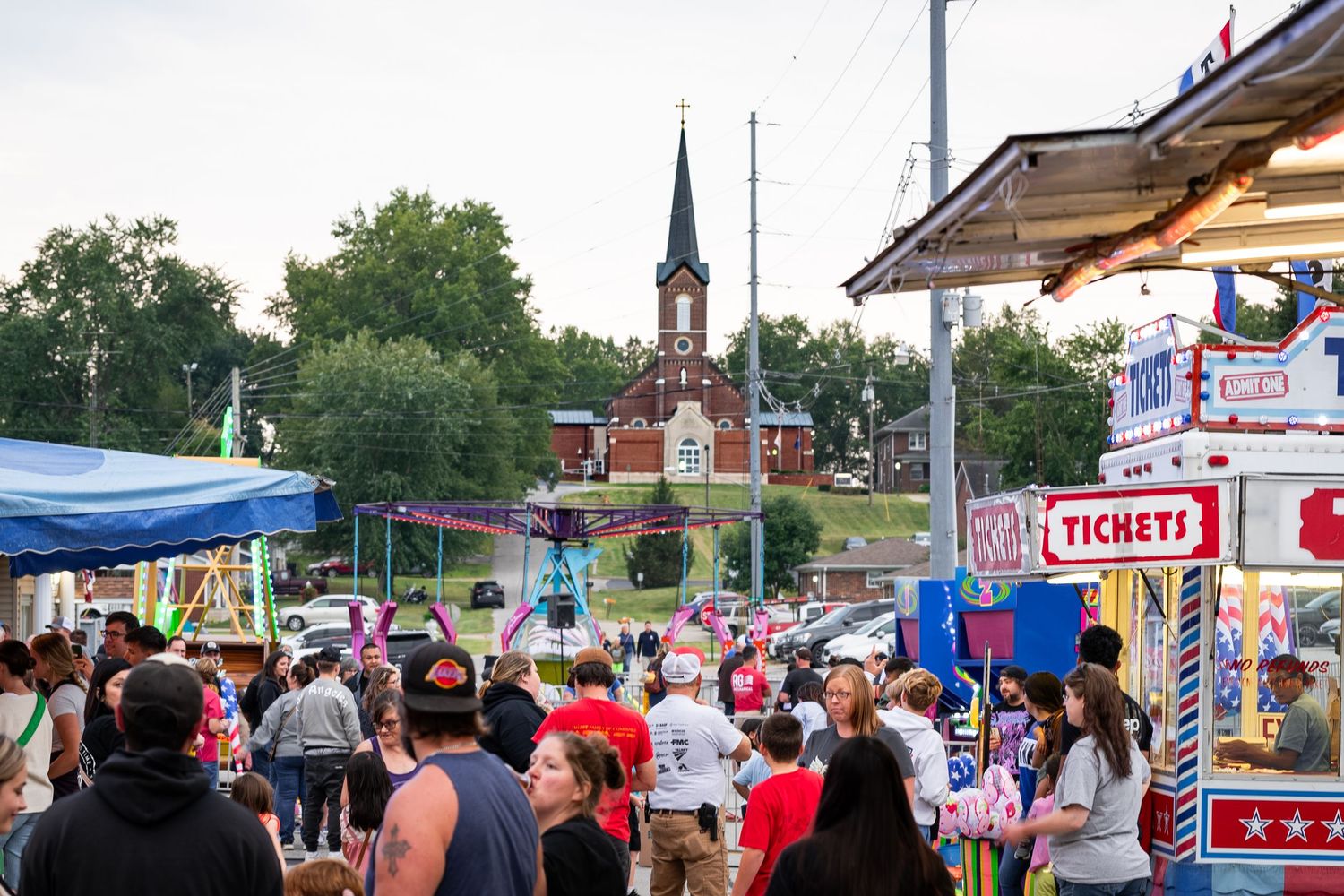 A lively outdoor carnival with a ticket booth, rides, and a church steeple visible in the background.