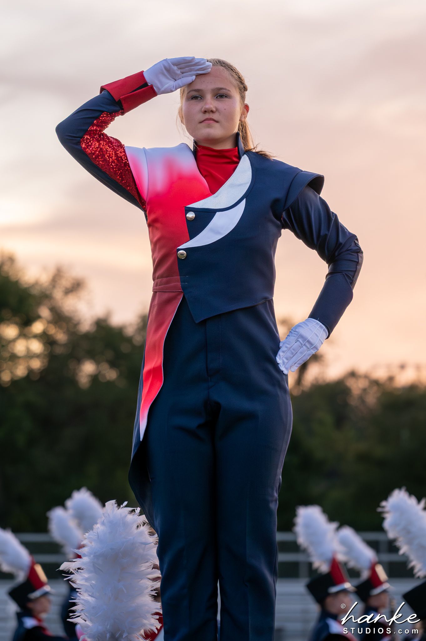A young female marching band member in a red and navy uniform salutes during an outdoor performance at sunset.