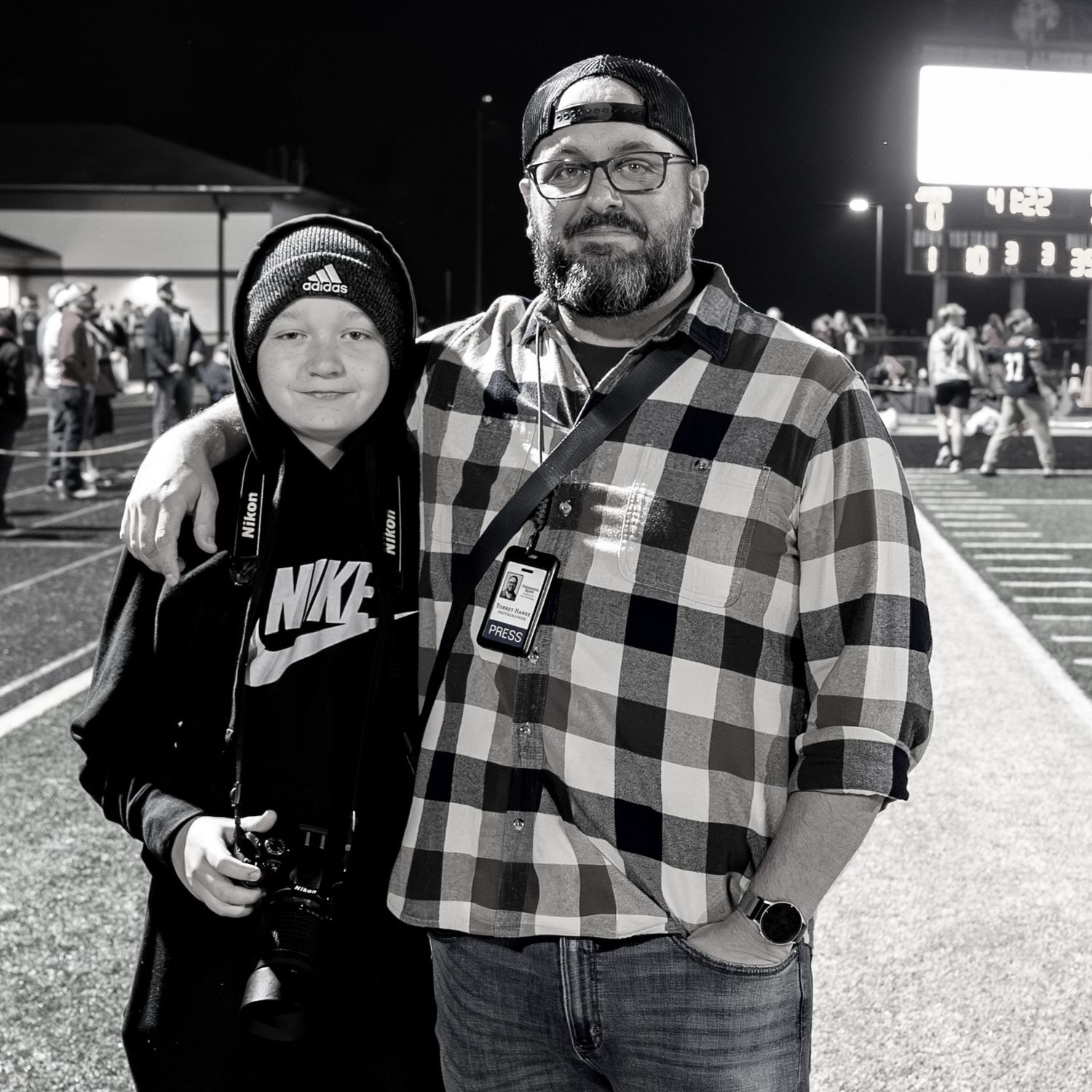 Black and white photo of Torrey Hanke, co owner of Hanke Studios, and his son posing together on a football field sideline.