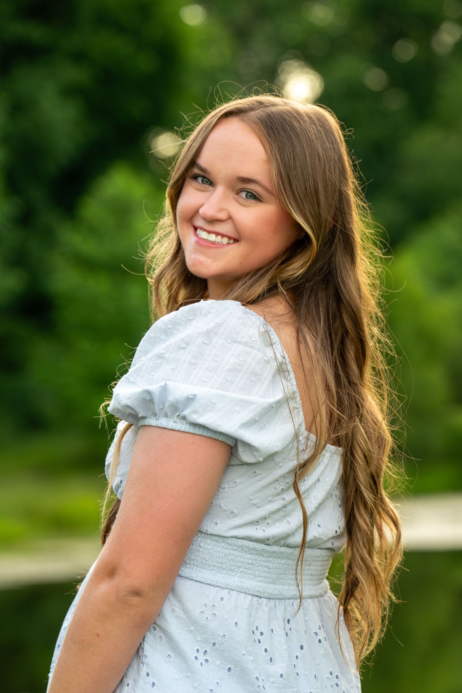 Young woman with long wavy hair smiling outdoors in a white dress, surrounded by lush green trees.