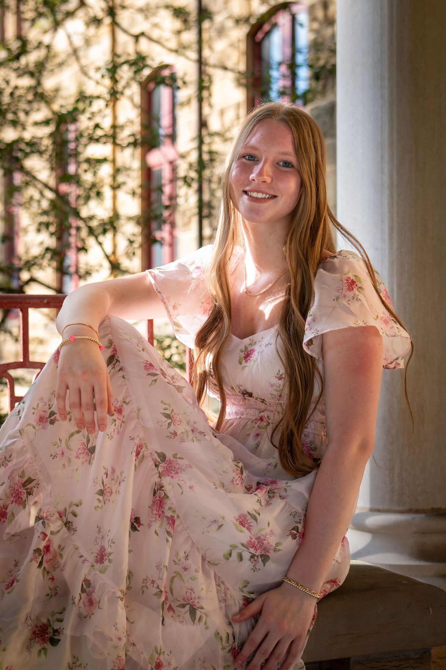 Young woman with long red hair smiling in a floral pink dress, seated on steps in a sunlit outdoor setting.