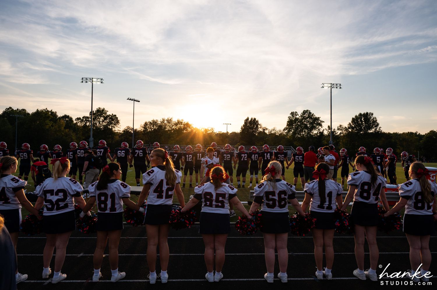 Cheerleaders and football players hold hands on the field at sunset during a high school game ceremony.