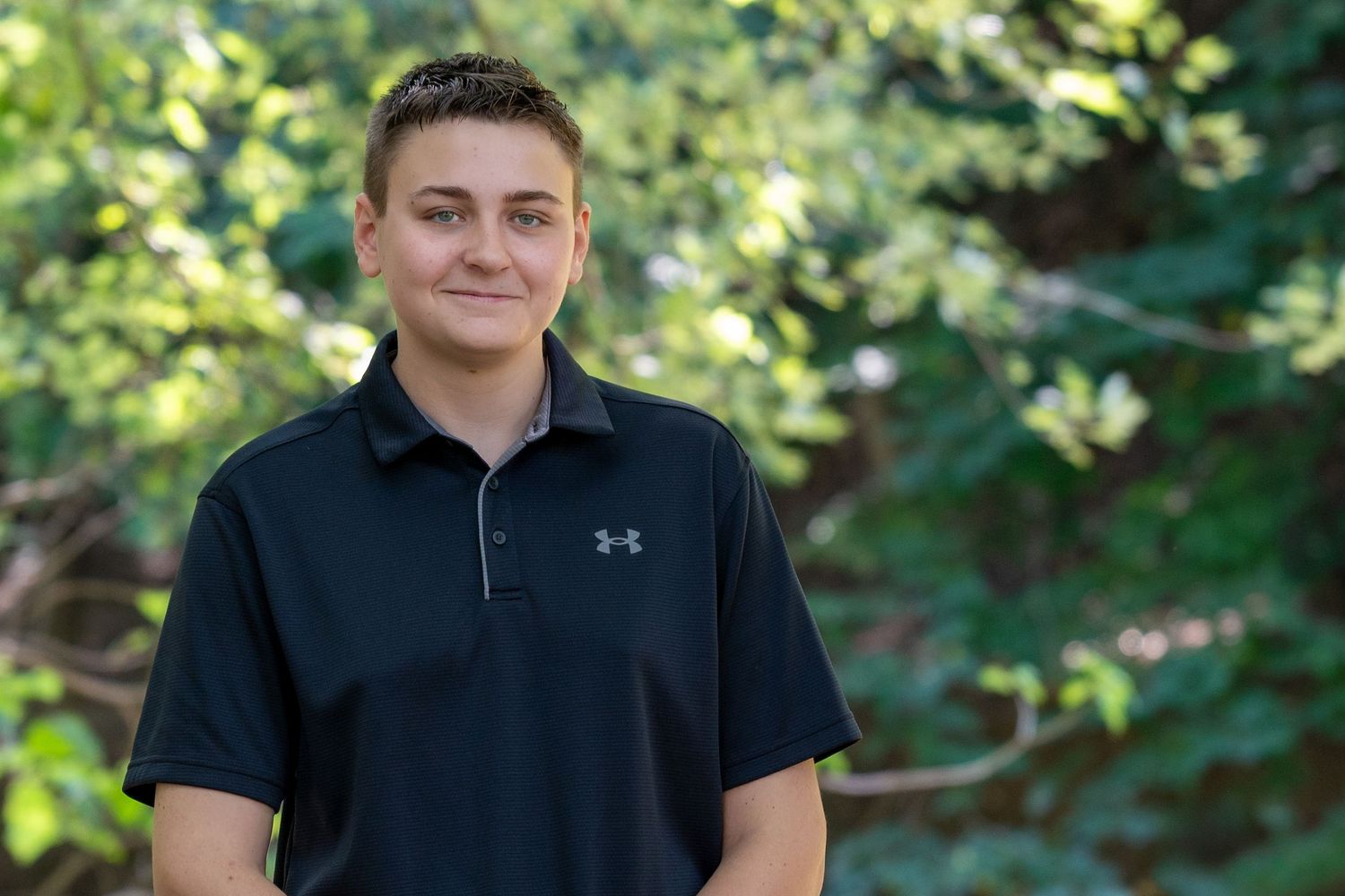 Young man in black Under Armour polo shirt smiling outdoors with green trees in background.