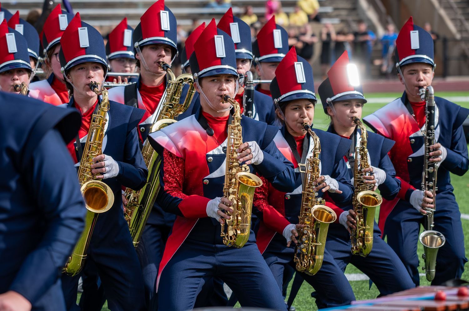Marching band members in red and navy uniforms play saxophones during a field performance at a stadium.