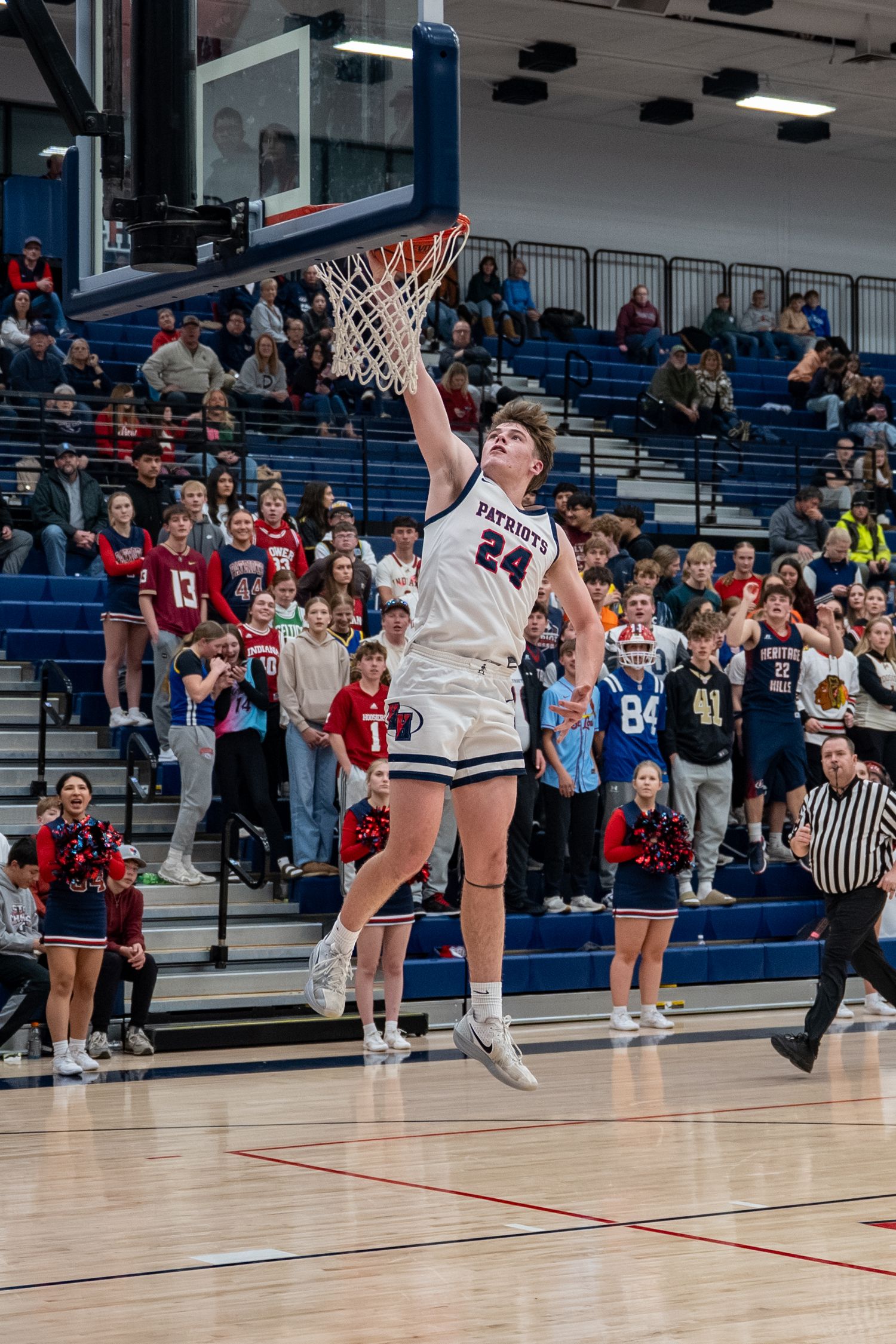 Player #24 in white Patriots jersey dunks basketball in packed high school gym during competitive game.